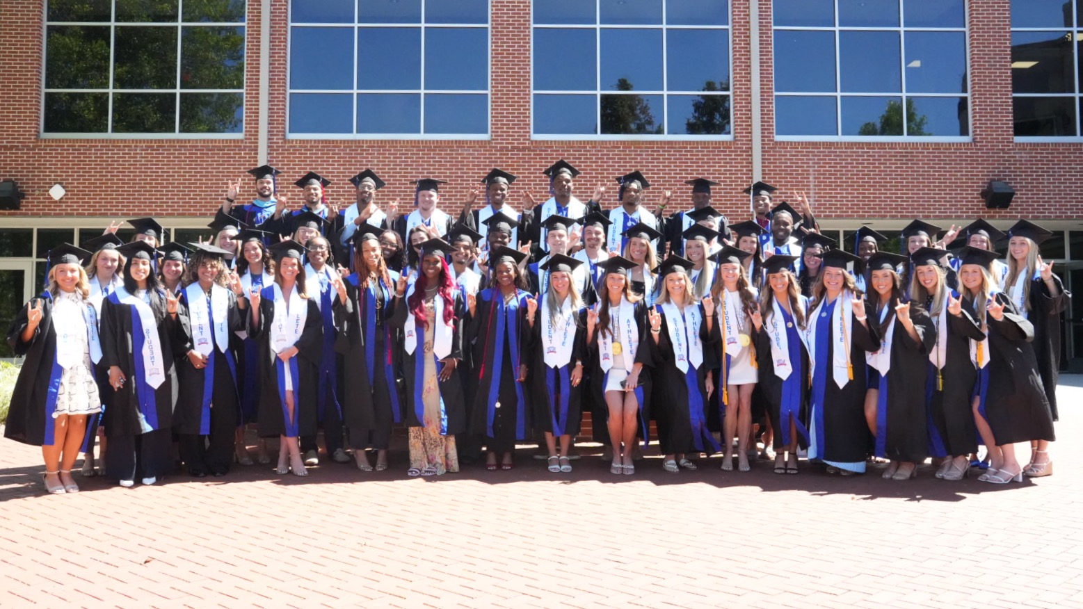 A group of UWG student-athletes and support staff pose holding the Wolf Hands sign while wearing their cap and gowns ahead of their 2026 commencement.