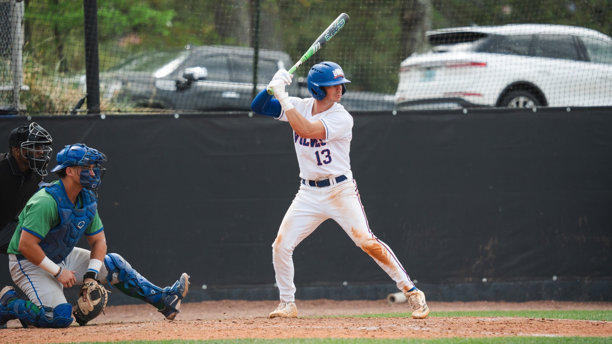 Baseball player in green jersey and blue gear kneeled down waiting to catch pitch. Player in white uniform with blue helmet next to him waiting to swing bat 