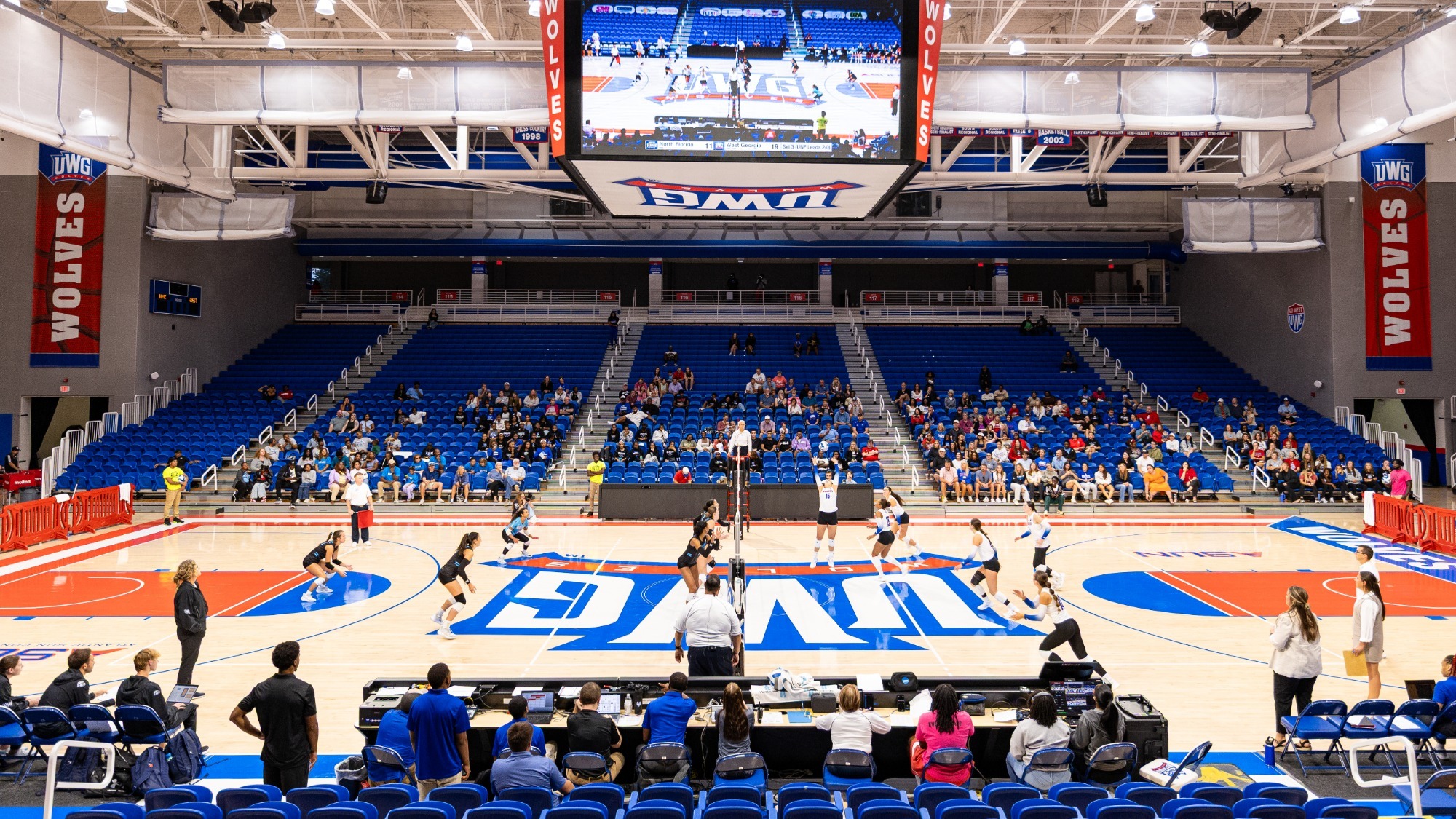 Pictured is the UWG volleyball court. Left side has a team in dark colors playing and the right team is in white. Fans in the background sitting in blue stadium seating 