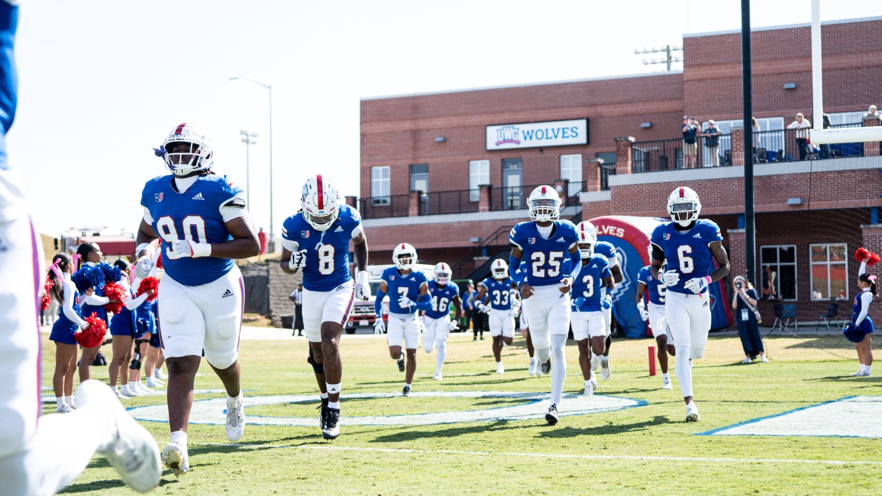 University of West Georgia football players in blue jerseys run onto the field before a game as cheerleaders line the entrance and spectators watch from a nearby building.