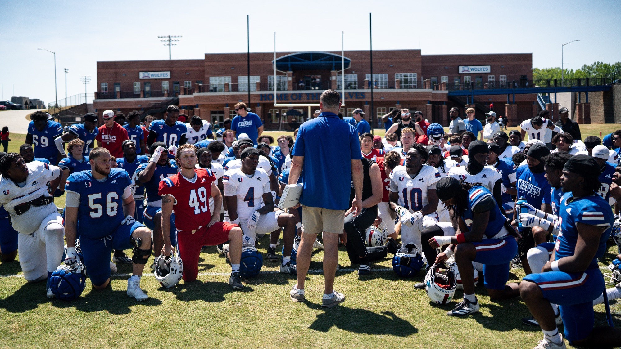 Head Coach Steve Englehart with Team huddled around him with the Athletics Operations Building behind him