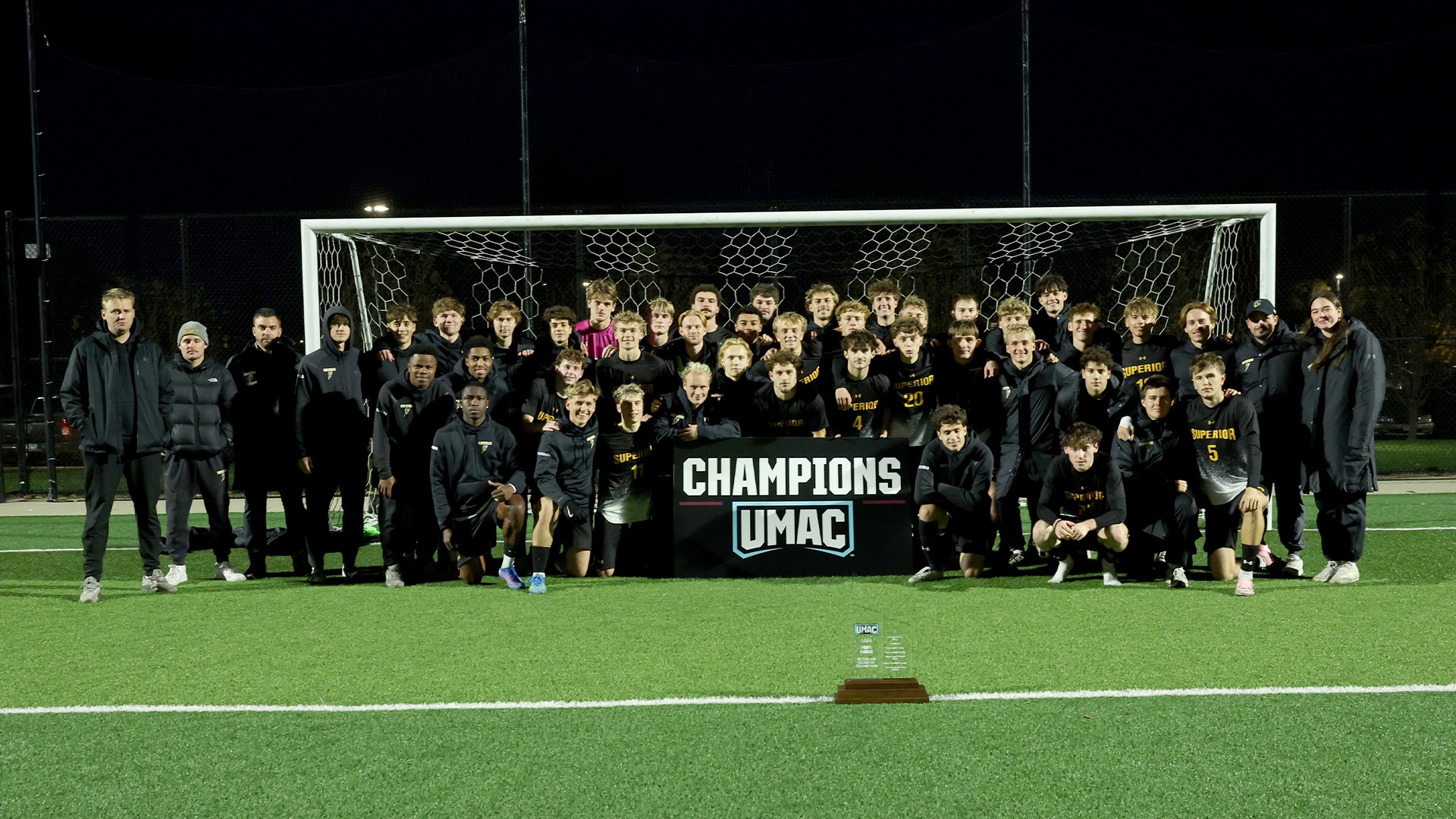 The Yellowjacket men's soccer team posing with a UMAC Champions banner in front of them along with a trophy a sizeable distance away.