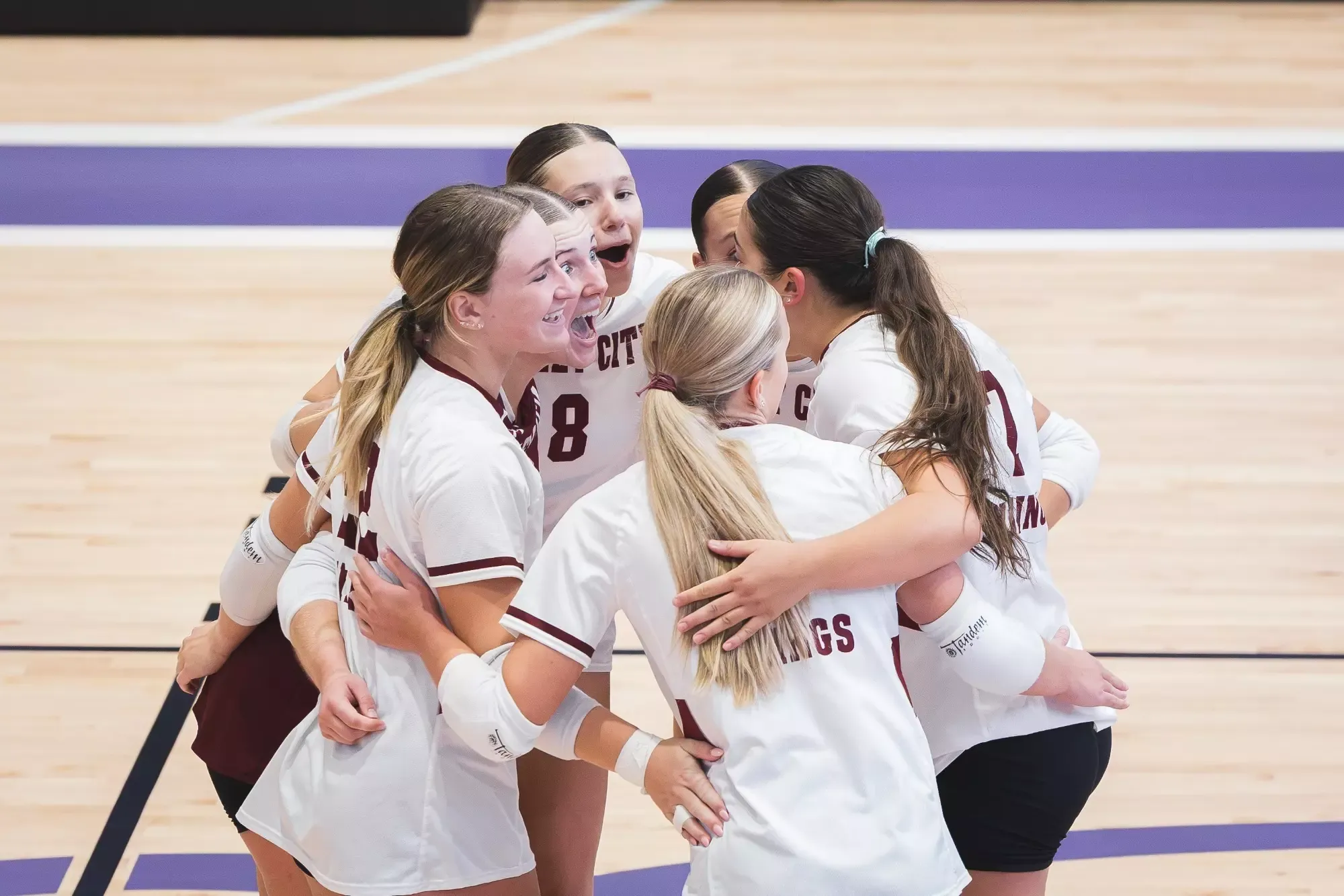 VCSU volleyball players in white uniforms embrace and celebrate together in a huddle on the court.