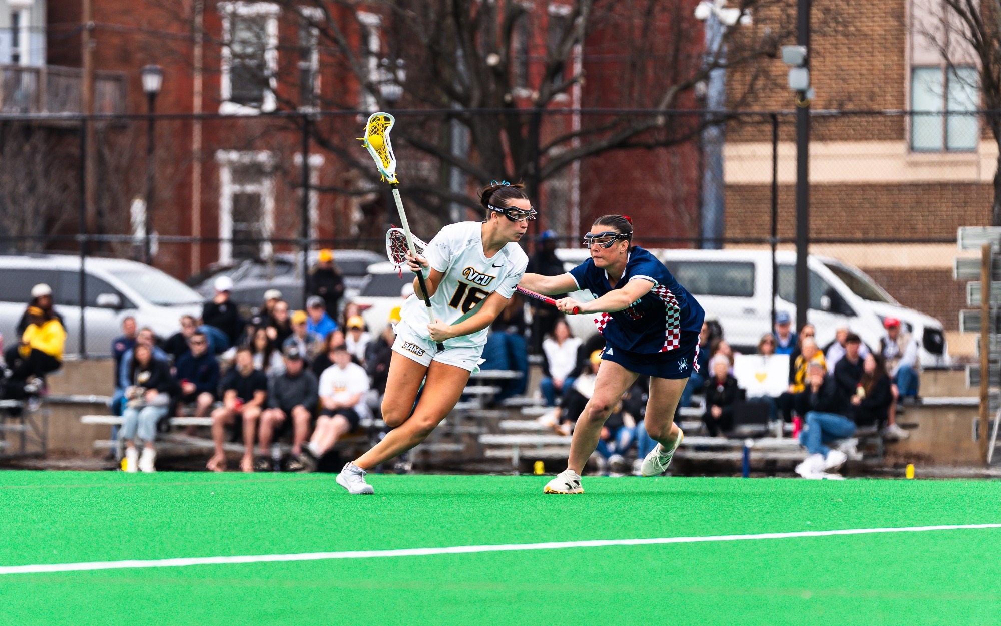 Kate Unzicker of VCU Lacrosse wearing a white 16 jersey runs with the ball while guarded by a Richmond defender