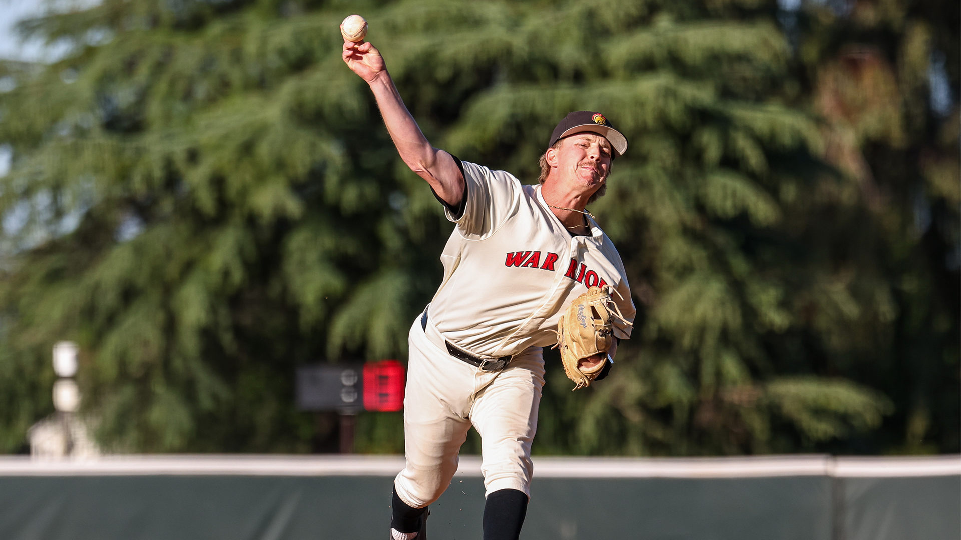 Evan Fanconi, wearing a light-colored Warriors baseball uniform and cap, delivers a pitch with his arm fully extended on an outdoor field, with a blurred backdrop of trees.