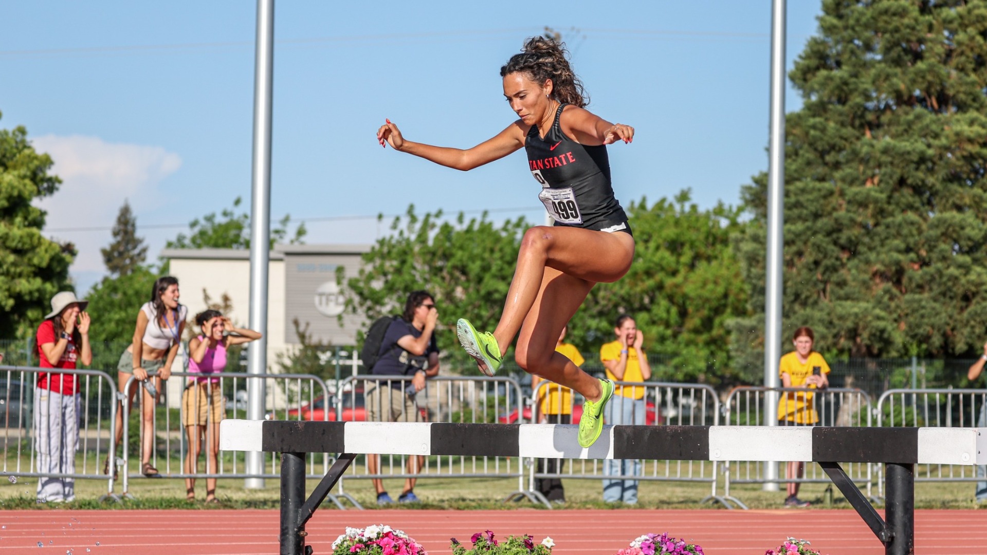 Photo of Stan State distance runner Bela Moreno clearing a steeplechase barrier over the water pit during a race, with spectators watching in the background.