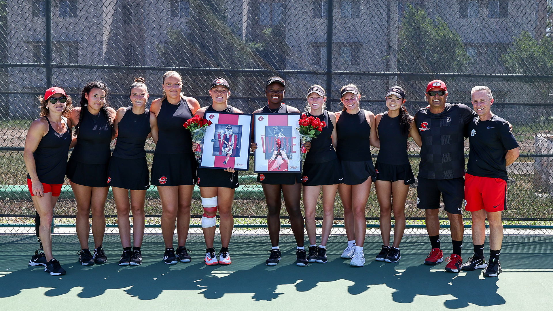 Stan State women’s tennis team and coaches pose on court for Senior Day, with two players holding framed posters and roses.