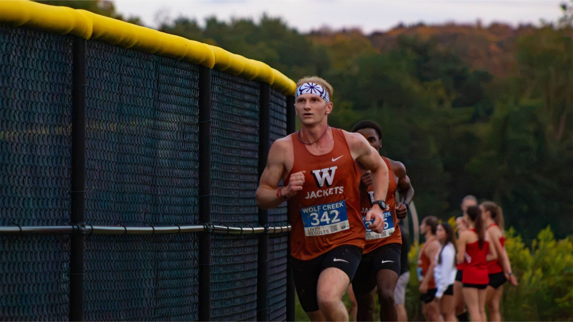A Waynesburg University men's cross country runner wearing a blue and white head band runs ahead of the field