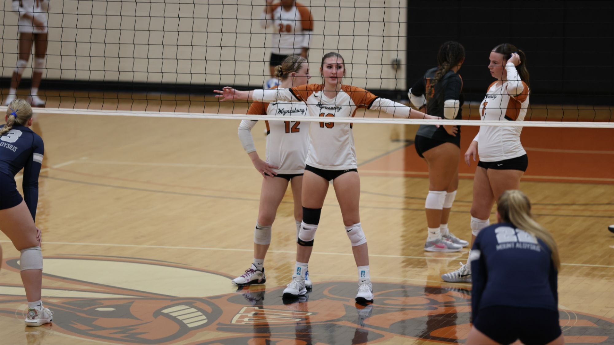 Izzy Shane stands near the net with her arms outstretched in front of the net while her teammates join her in waiting for the serve