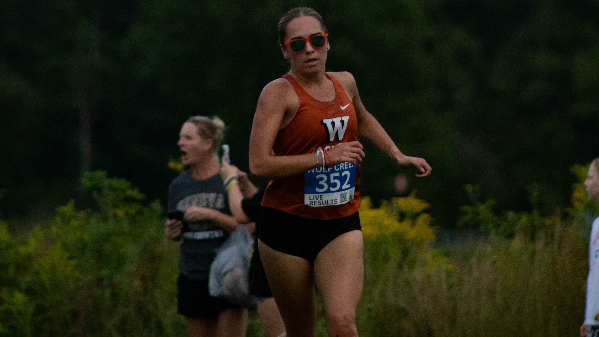 A Waynesburg University women's cross country runner wearing black sunglasses runs on the course