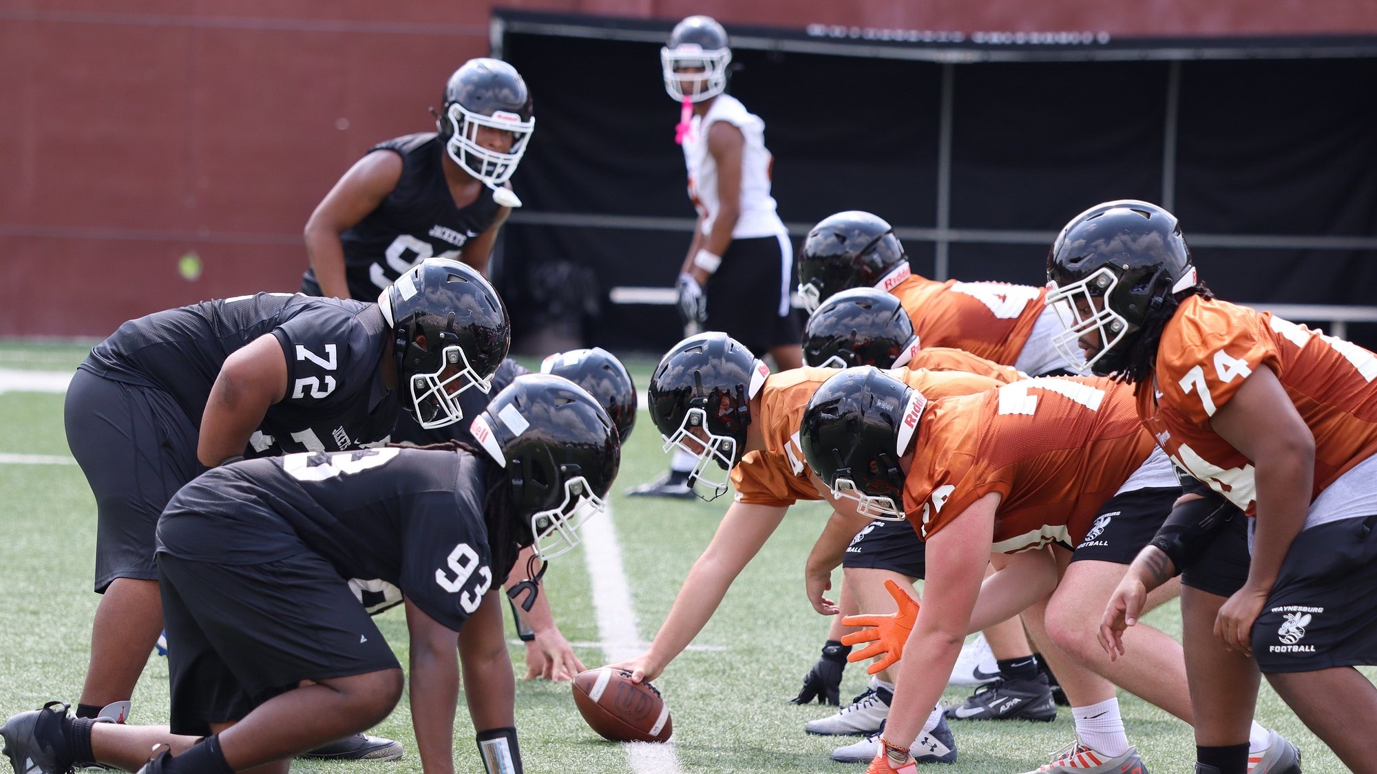 The Waynesburg defensive line, wearing black jerseys, and the offensive line, wearing orange jerseys, line up across each other in training camp