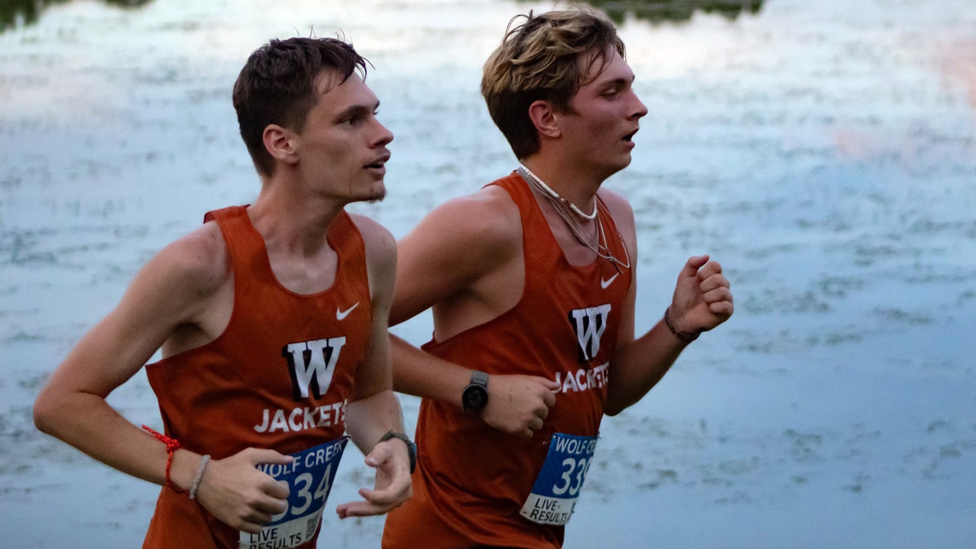 Two Waynesburg University men's cross country runners in orange running jerseys run in front of a body of water