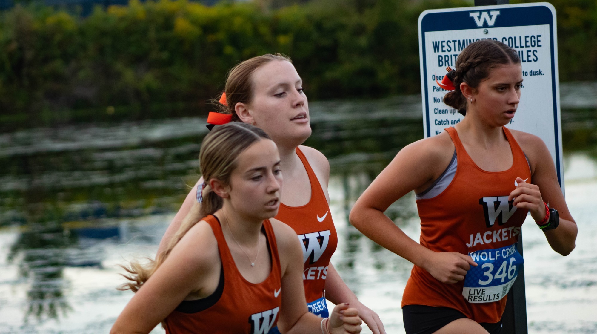 Three Waynesburg University women's runners in orange uniforms run in front of a sign and a body of water