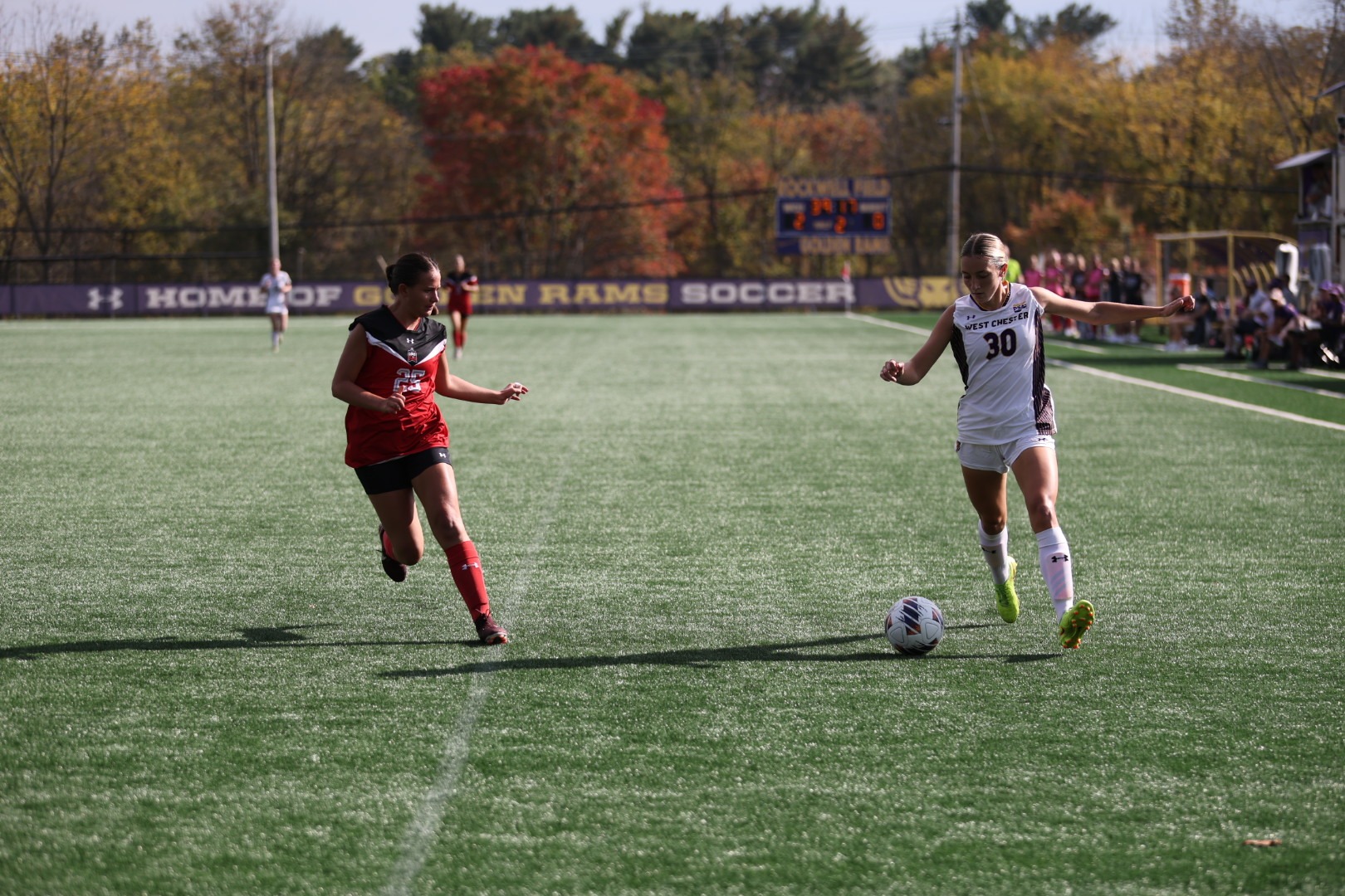 Lindsay Balmer wearing home white on white kit carries the ball down the wing vs East Stroudsburg with an ESU defender wearing red and black jersey with black shorts closing the gap