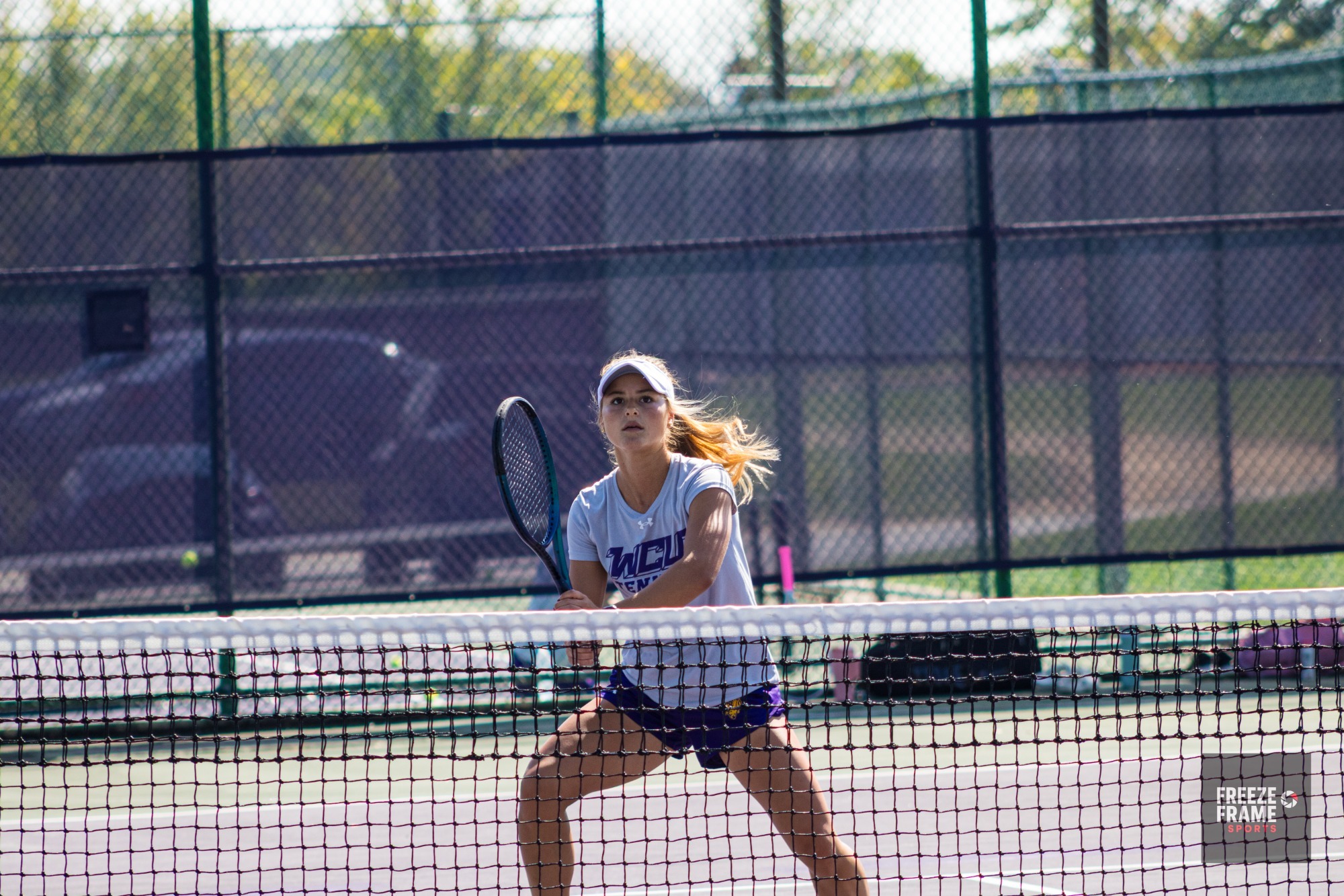 WOMEN'S TENNIS PLAYER WEARING GRAY T SHIRT, VISOR AND PURPLE SHORTS IN FRONT OF THE NET TRACKING BALL