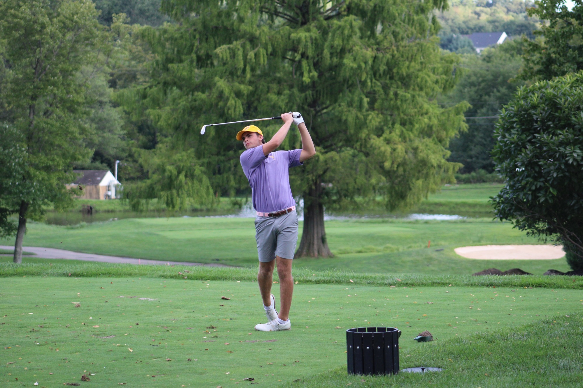 Will Hilton wearing purple polo, gray shorts, yellow hat, and white shoes holding follow through after teeing off on a par three at penn oaks in west chester