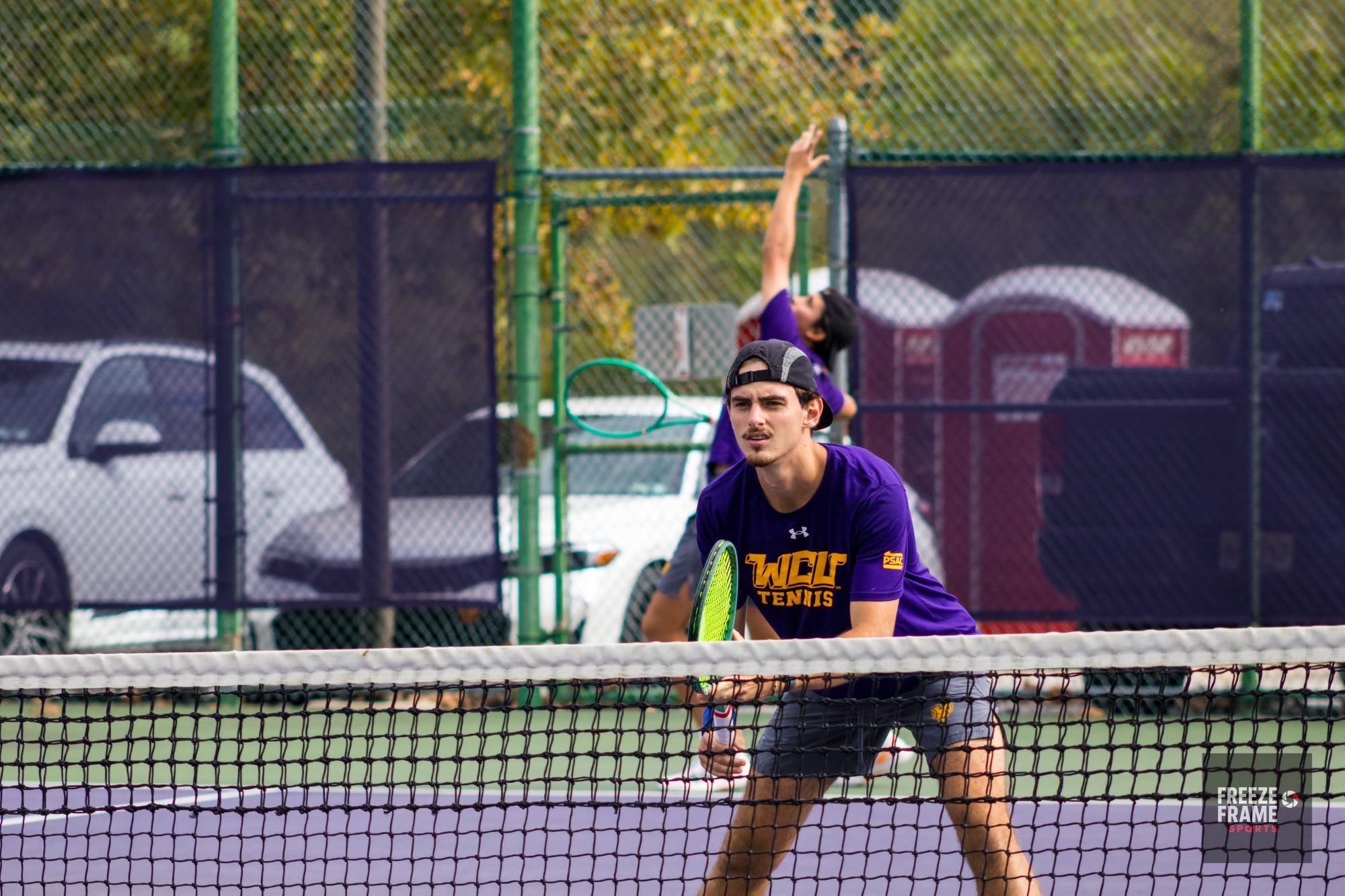 Thomaz Joly close to net with backwards hat and racket facing net with JP Beirana Guzman preparing to serve behind him