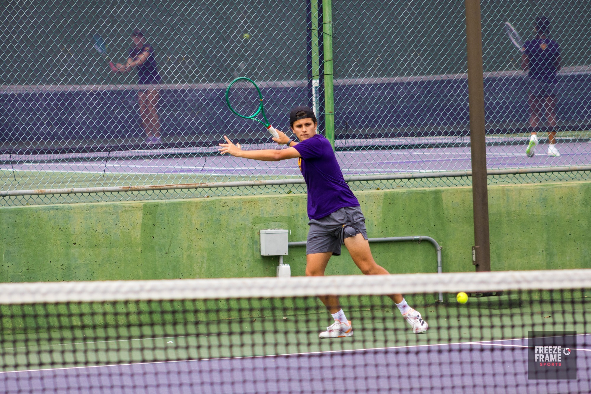 MEN'S TENNIS PLAYER PLAYING A FOREHAND SHOT WEARING PURPLE SHIRT IN FRONT OF PURPLE WINDSCREENS