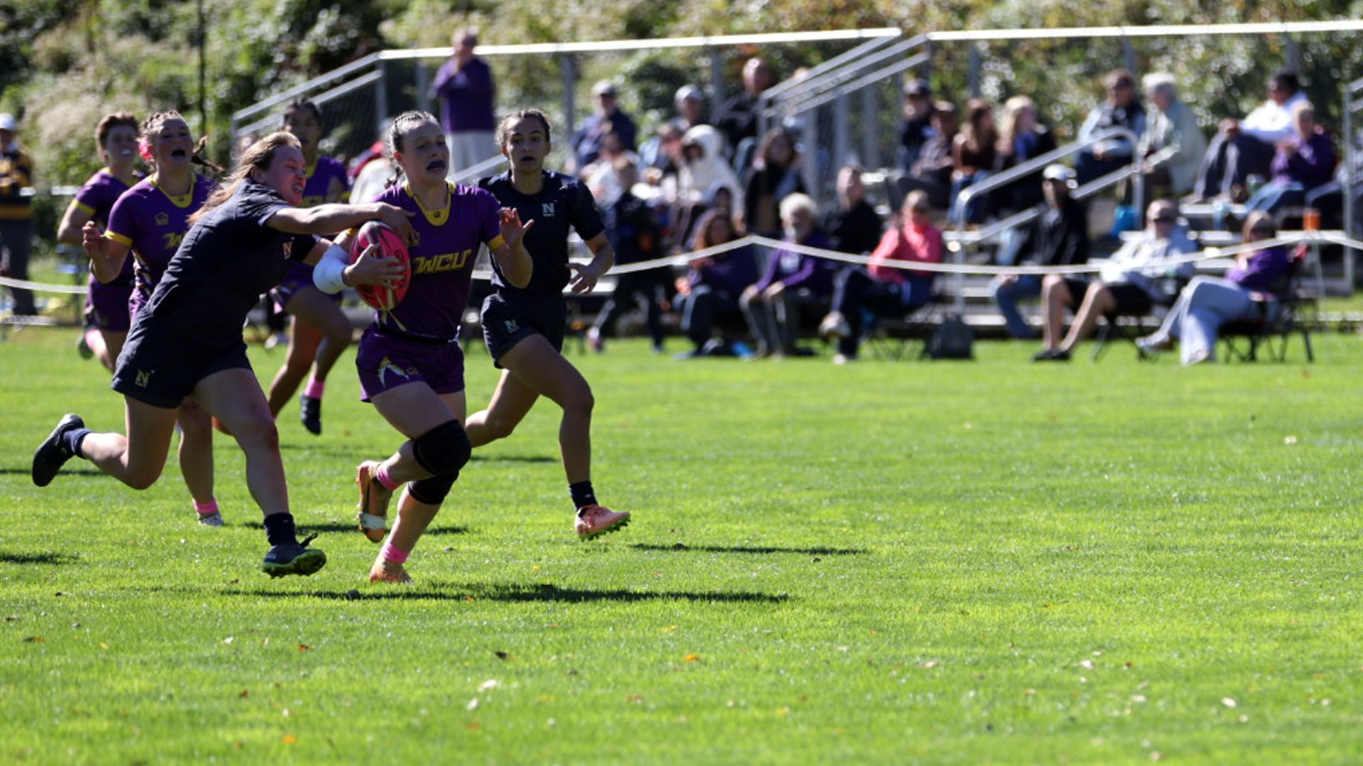 WCU WOMEN'S RUGBY PLAYER WEARING PURPLE AND GOLD CARRIES HTE BALL UP FIELD VS NAVY WITH NAVAL ACADEMY PLAYER GIVING CHASE AND REACHING AROUND FOR BALL ON 10-18