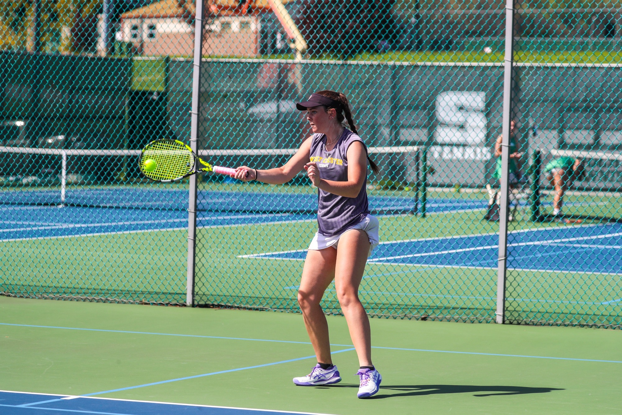 Brigitte Gravelle wearing black visor with Ram head logo, purple WCU tennis shirt, and white skirt with tennis ball rolled into side forehand at  SlipperyRock in 2026 PSAC Quarterfinals
