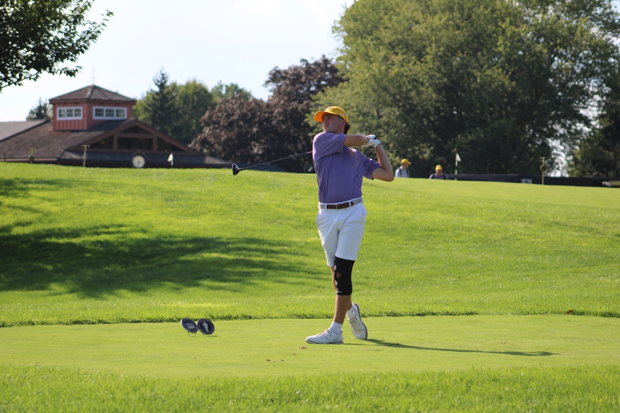 Nathan Radwanski wearing purple polo, yellow hat, white shorts and knee brace holding his follow through after a tee shoton tee box at Penn Oaks
