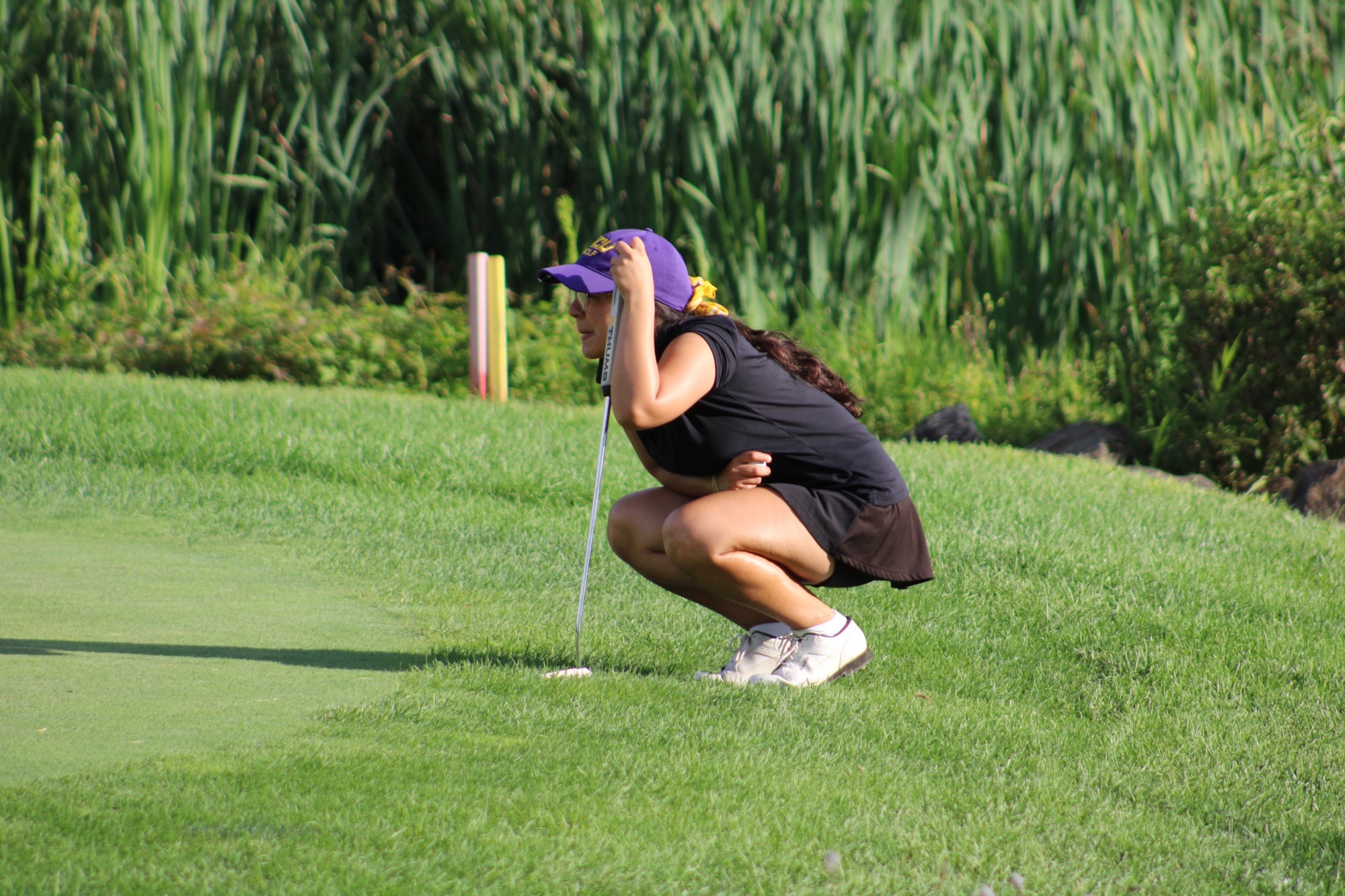 Fatima Rizvi wearing black polo, black skirt, purple hat with gold bow crouches off the green to read a putt with putt in hand at penn oaks