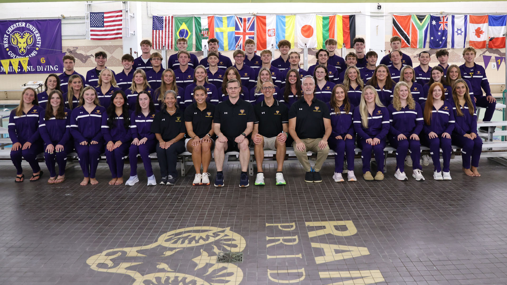 Men's and Women's Swimming & Diving Team Photo with both teams together on the deck of Graham Natatorium's pool. Athletes in their purple warmup suits and coaches in their black polo shirts