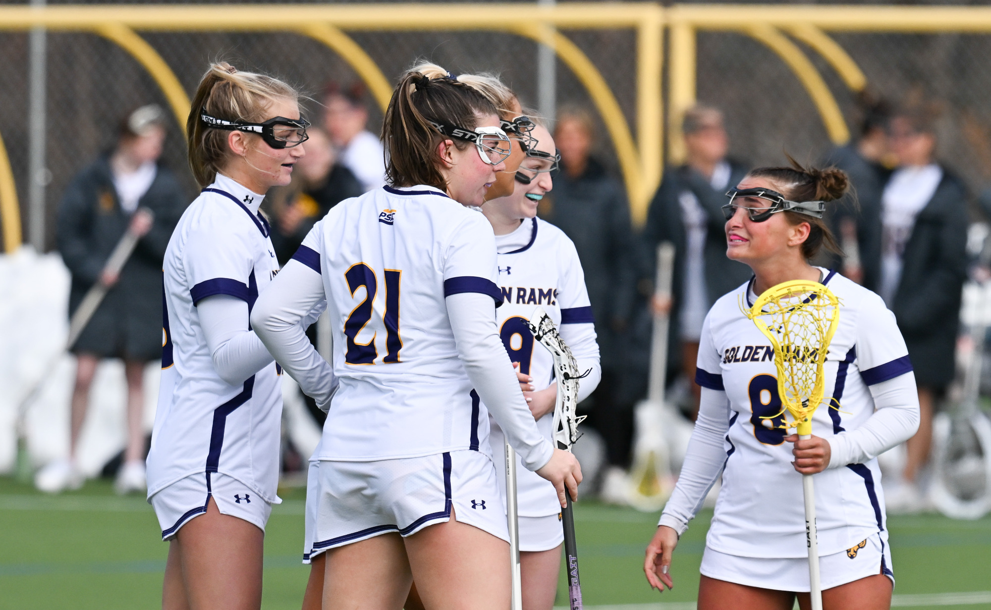 Three Women's Lacrosse players in their home white uniform with purple trim celebrate a goal in season opener at Vonnie Gros Field