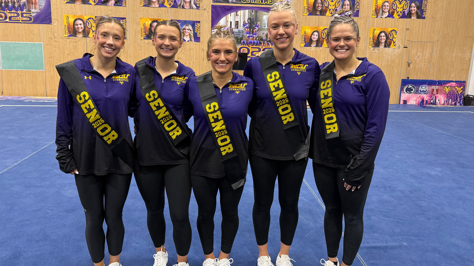 Women's Gymnastics Seniors Women's Gymnastics seniors pose for a photo on Senior Day with their coaches wearing purple warm up top with a sash arond right shoulder that says Seniors and black leggings with white sneakers. From left to right Melina Ringas, Sarah Eskew, Anna Lacey, Karah Vargo and Macarthy Keane.