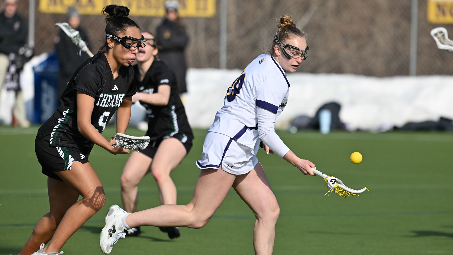 Kendall Fortune picks up a ground ball, with a Slippery Rock defender right on her back, dressed in WCU's home white jerseys with purple trim and purple numbers and Slippery Rock defender in her road black uniforms with white numbers in green trim.