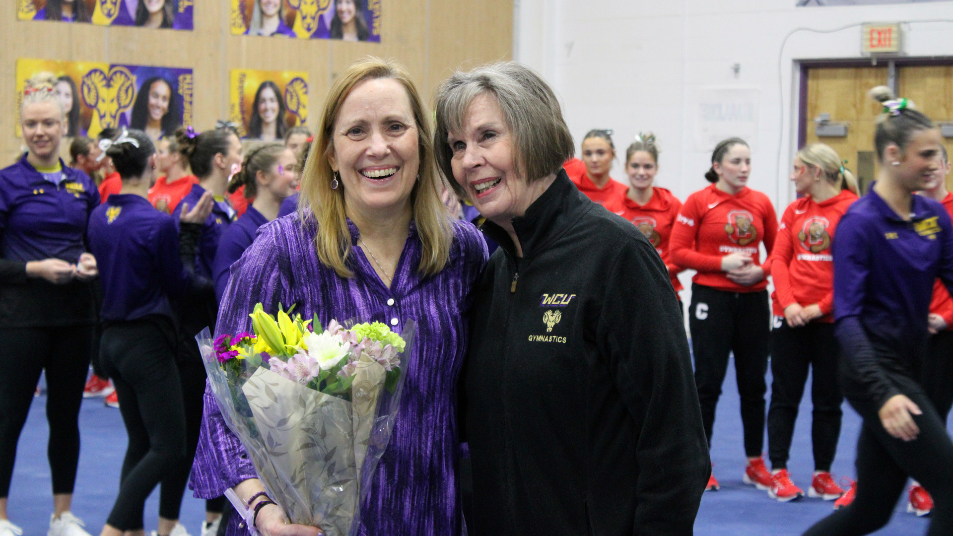 Barb Cordova and Sandy Thielz pose for a photo after Barb tied Sandy's career wins record. Barb is holding a bouquet of flowers, smiling, wearing a purple top while Sandy is in a black quarter zip fleece with WCU Gymnastics on the left side.