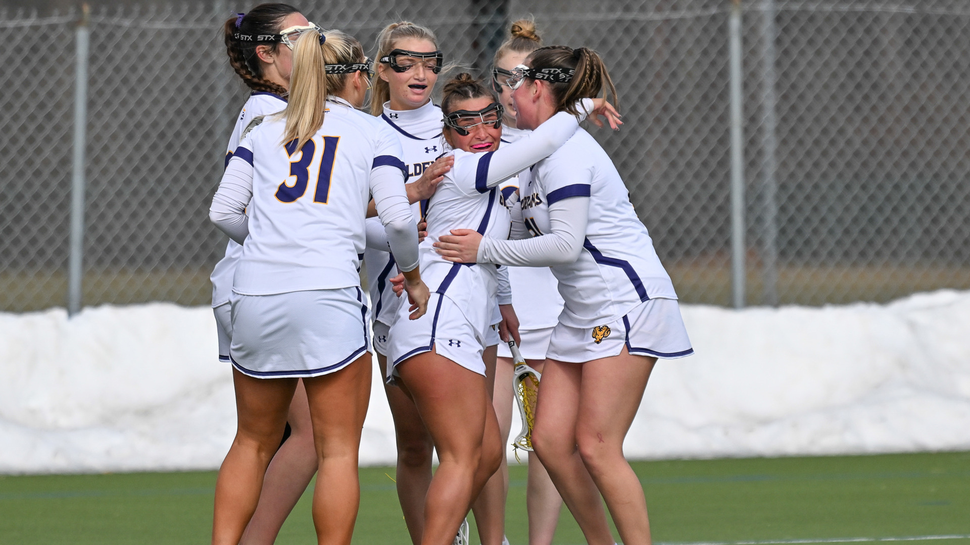 Six Women's Lacrosse players hug in celebration of a goal at home in their white jerseys with purple trim and letters