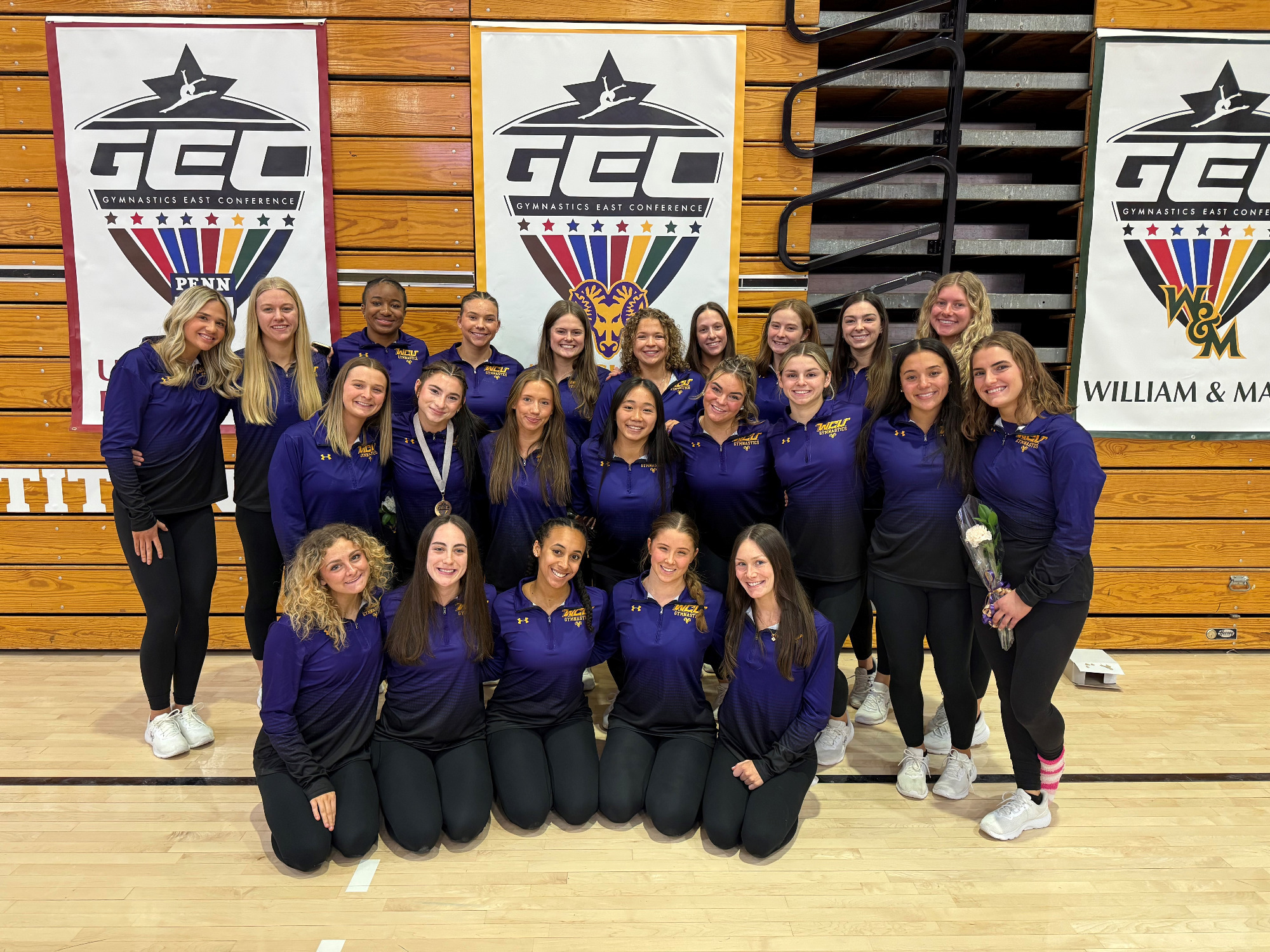 Women's Gymnastics Team at GEC Championships posed for team photo in their purple warmup suits; seniors holding flowers and Gianna Smith wearing her second place medal around her neck
