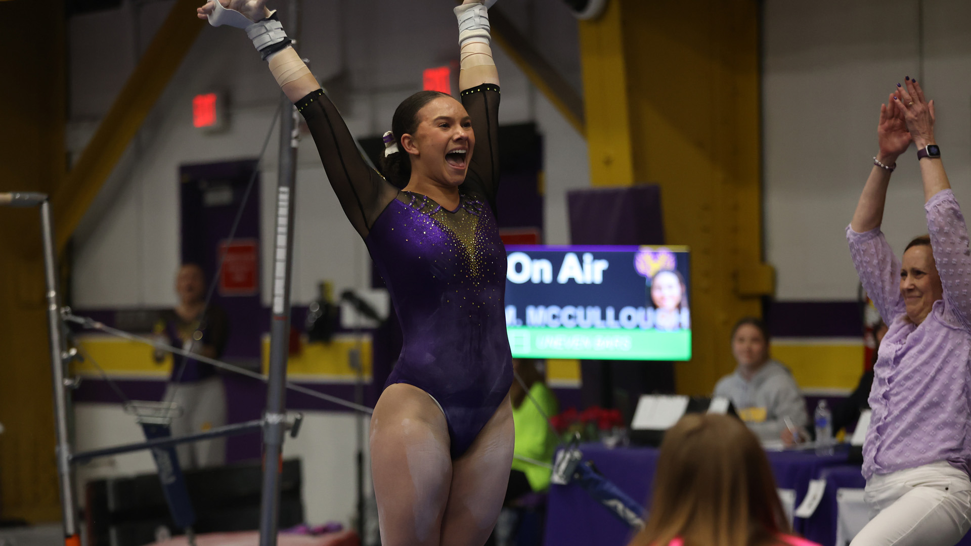 Madelyn McCullough smiles with hands raised in the air after her dismount from the uneven bars while head coach Barb Cordova claps emfatically
