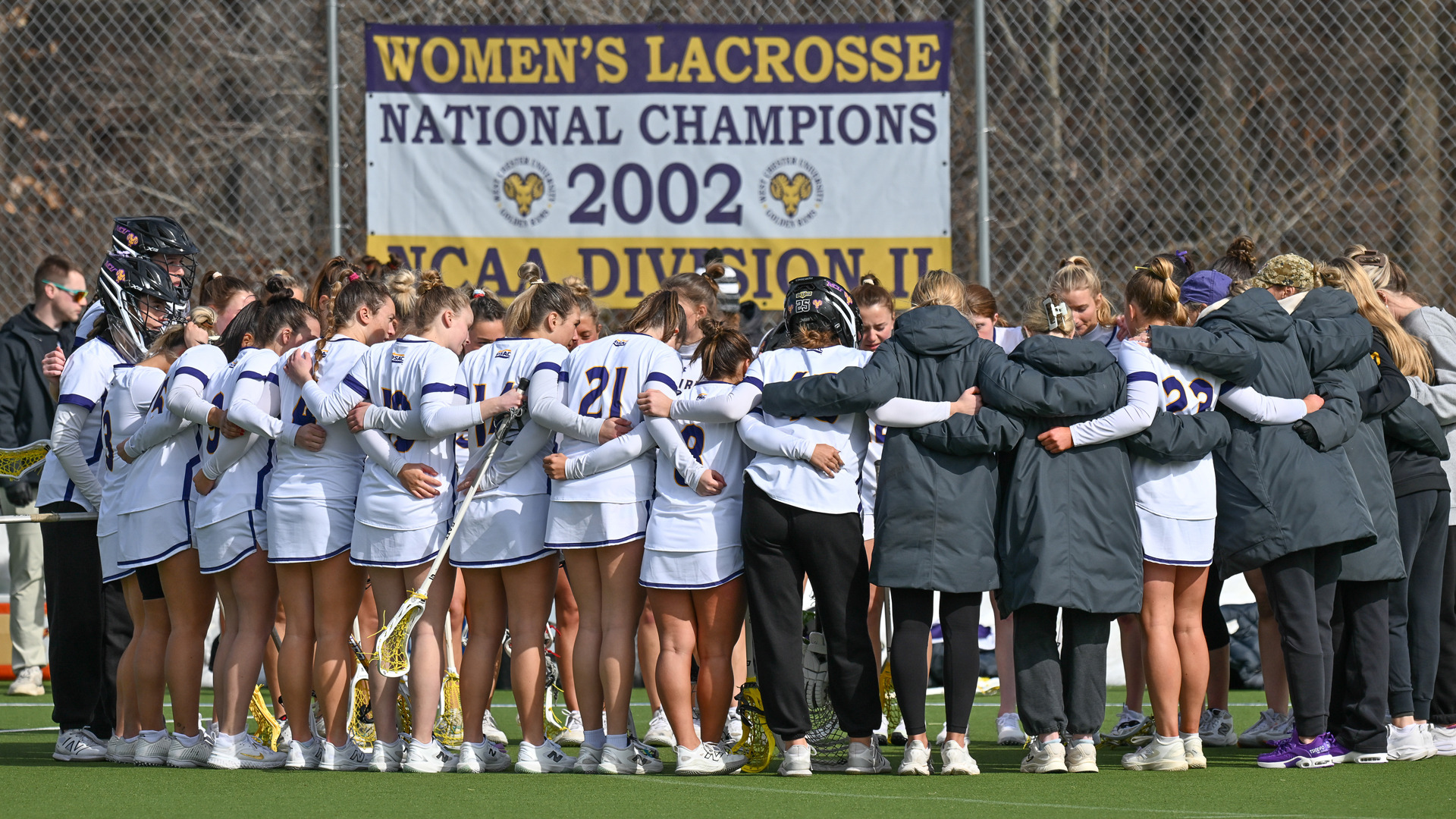 Women's Lacrosse squad in team huddle on sideline in the pregame wearing their home white jerseys with purple trim and purple West Chester on the front with purple numbers and some players wearing winter puffer jackets to keep warm.