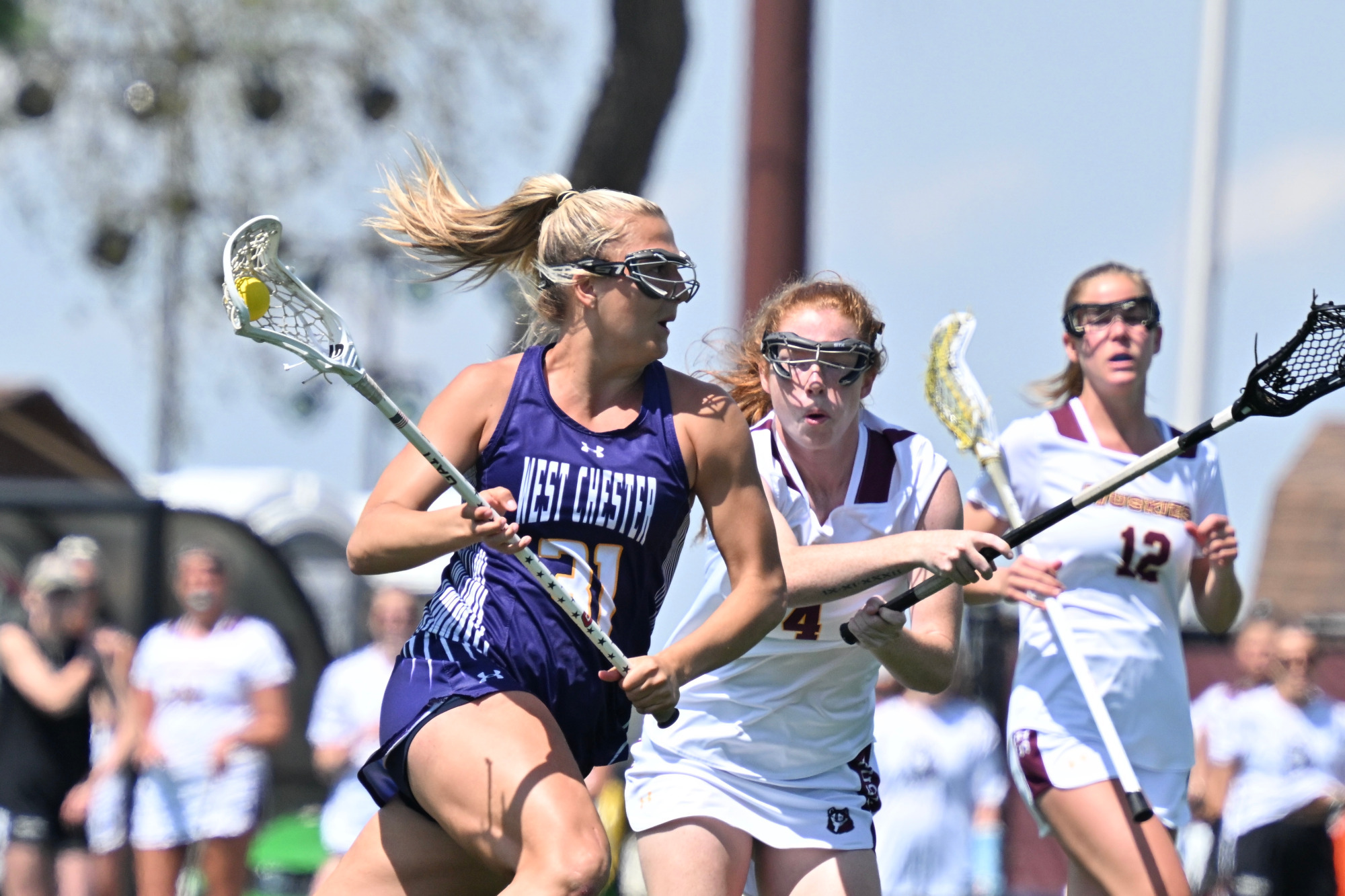 Sydney Wasdick races around a Bloomsburg defender while cradling the ball in her purple road jersey with white numbers and yellow trim.