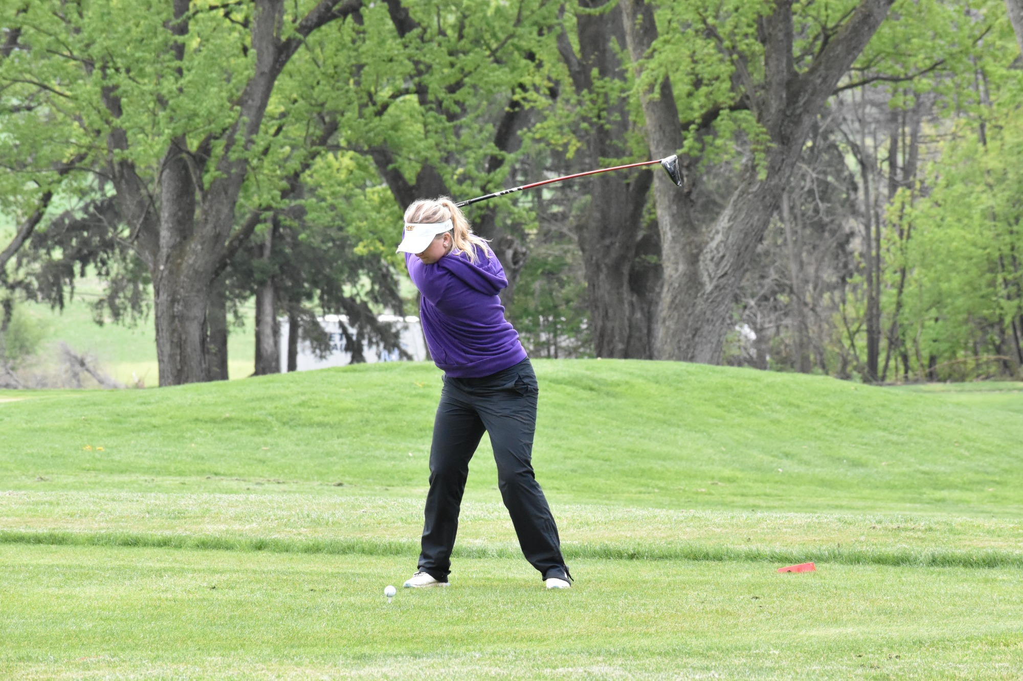 Hanna Lignell tees off on the first hole at the PSAC Women's Golf Championships on Wednesday wearing purple sweatshirt with Golden Ram logo, white hat, and black rainpants