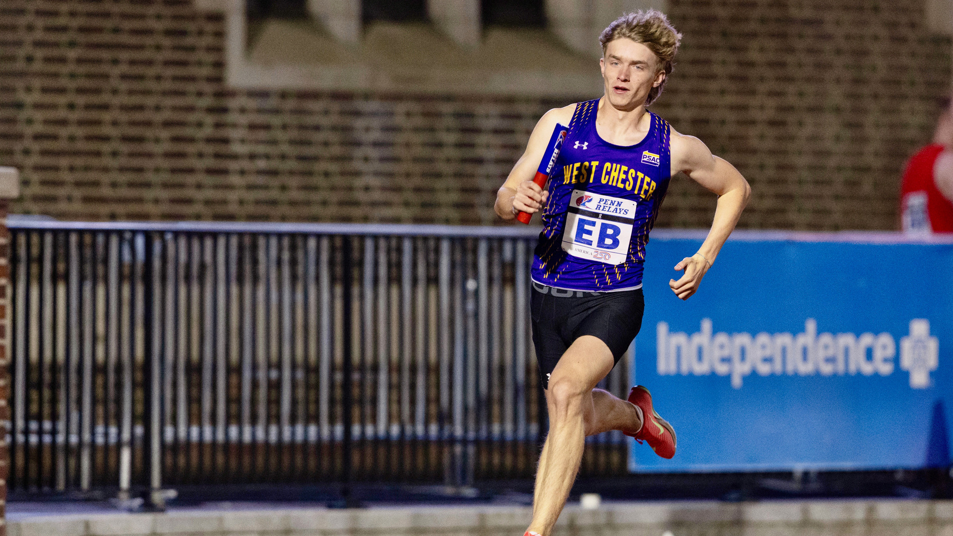 Jordan Barge comes around Turn 4 carrying the baton at the Penn Relays in his purple track uniform, black running shorts and red NIKE running shoes