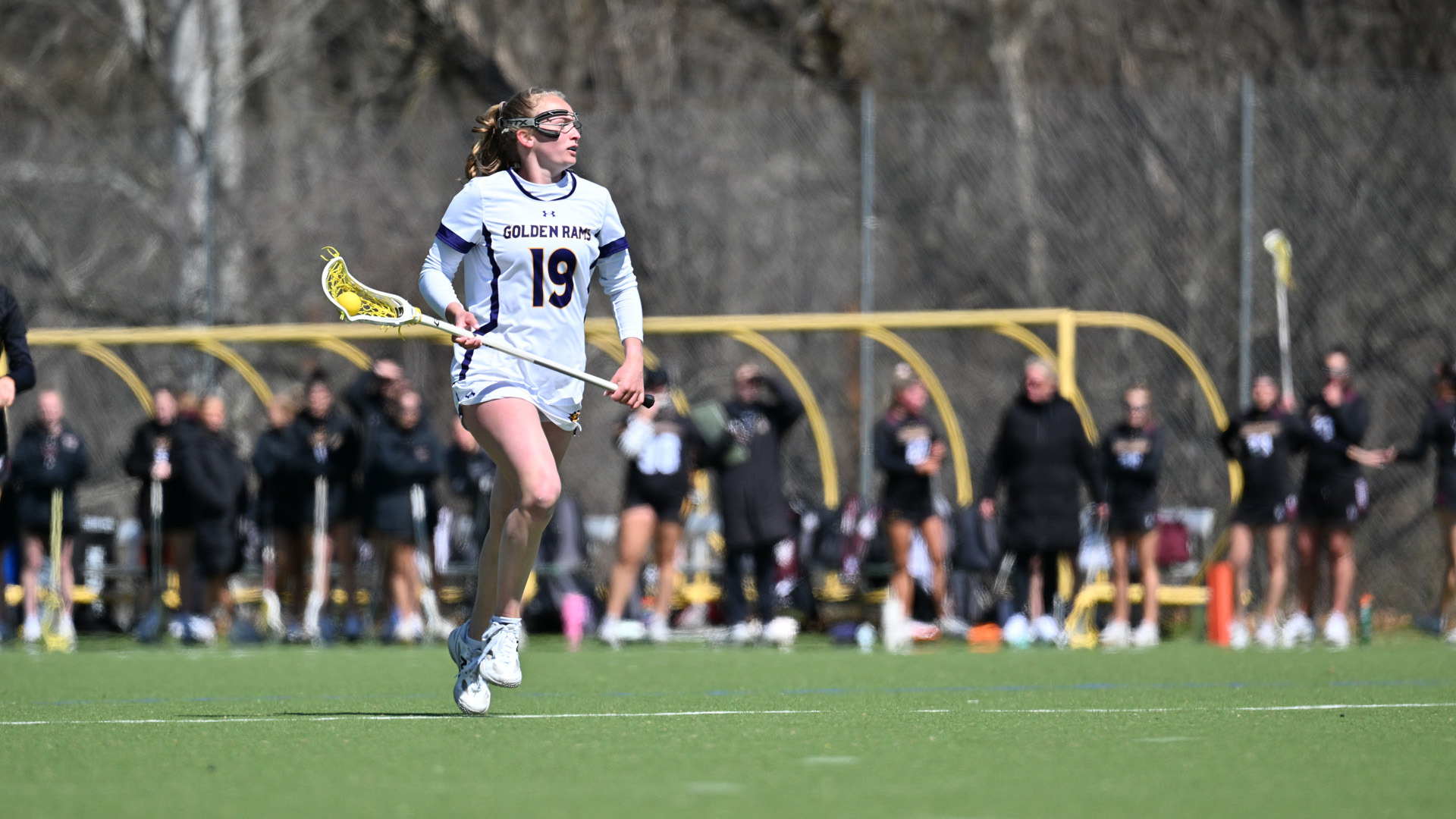 Kendall Fortune carries the ball over the restraining line against Bloomsburg in her white home jersey with purple numbers and letters in gold trim