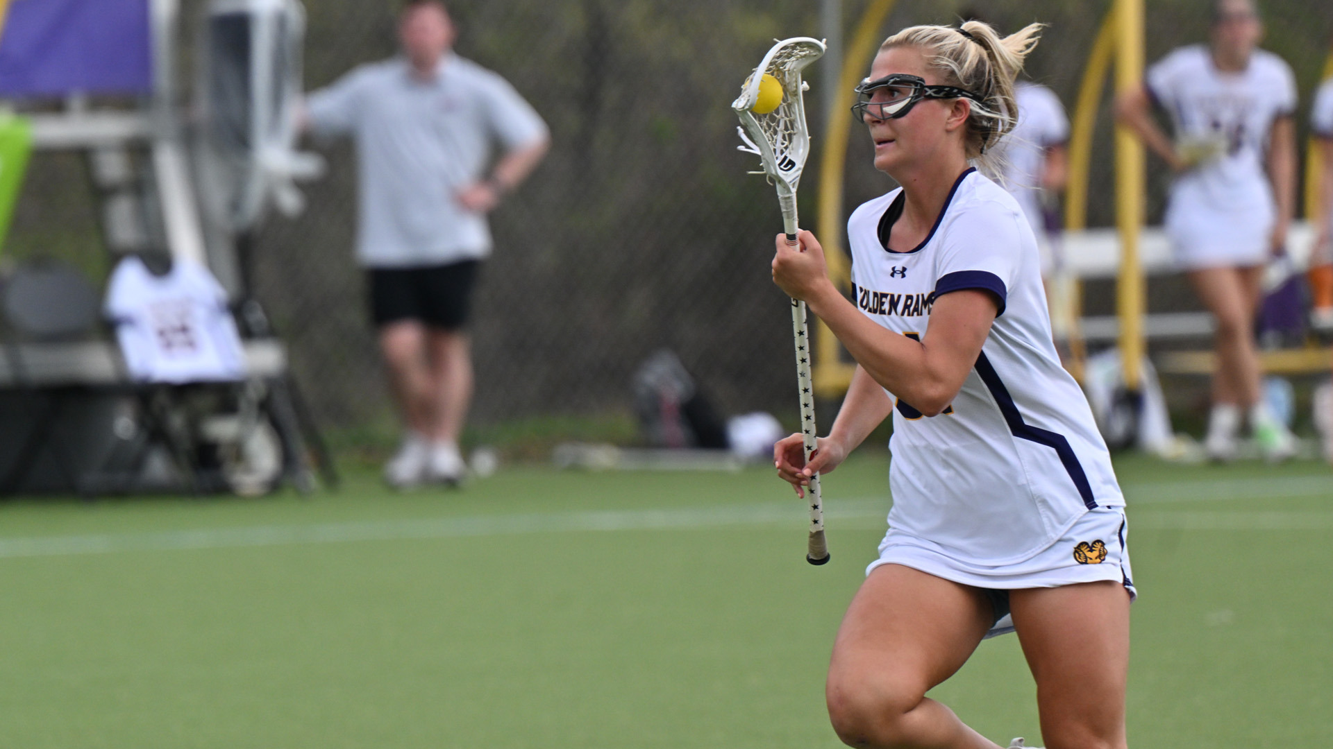 Sydney Wasdick races through the midfield at Vonnie Gros Field in her white home uniform with purple numbers and letters in gold trim against Bloomsburg