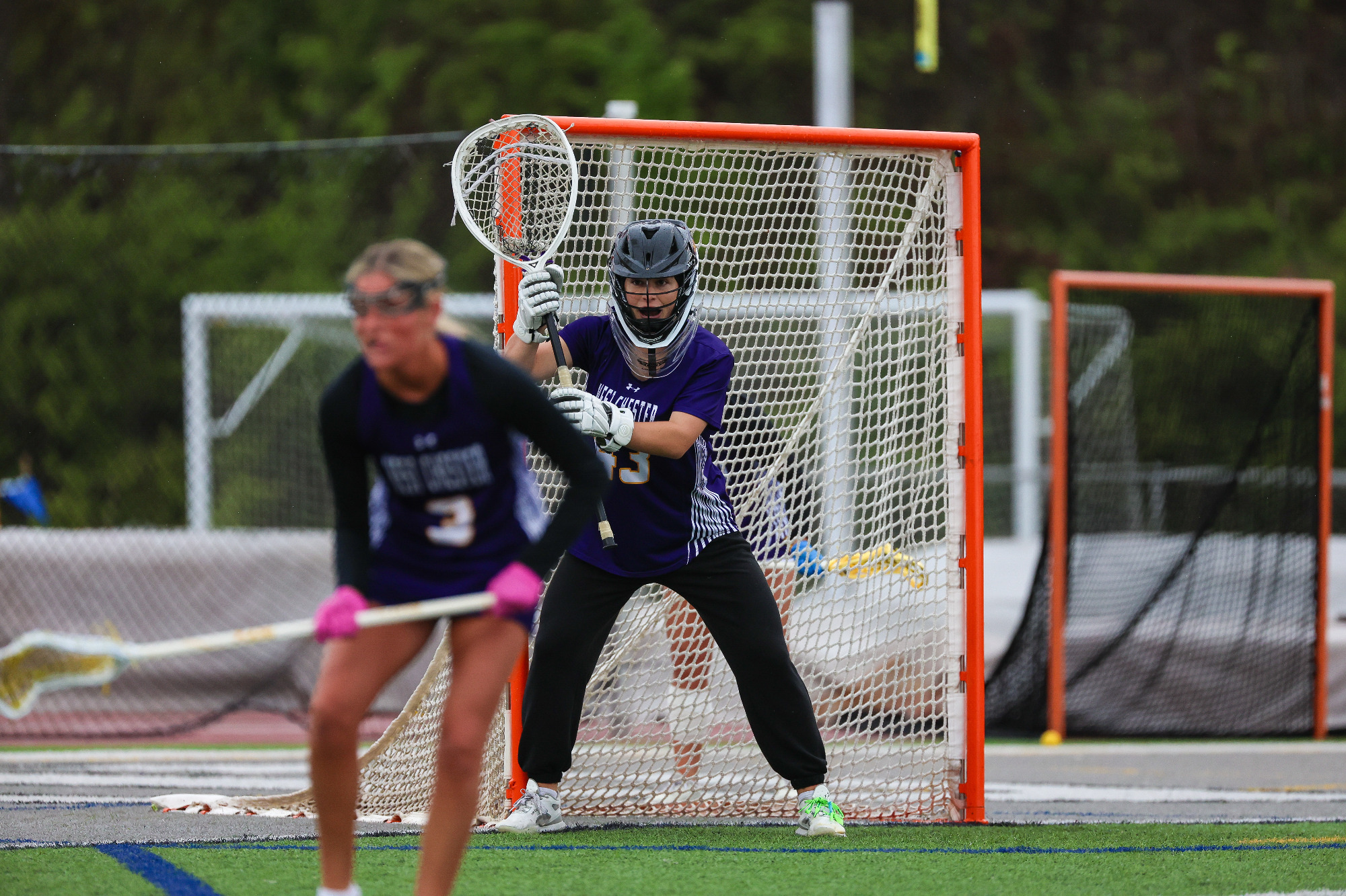 Morgan Koons stands ready for a shot in the cage in her road purple jersey and black sweatpants with defender Dani Bruno in front of her.