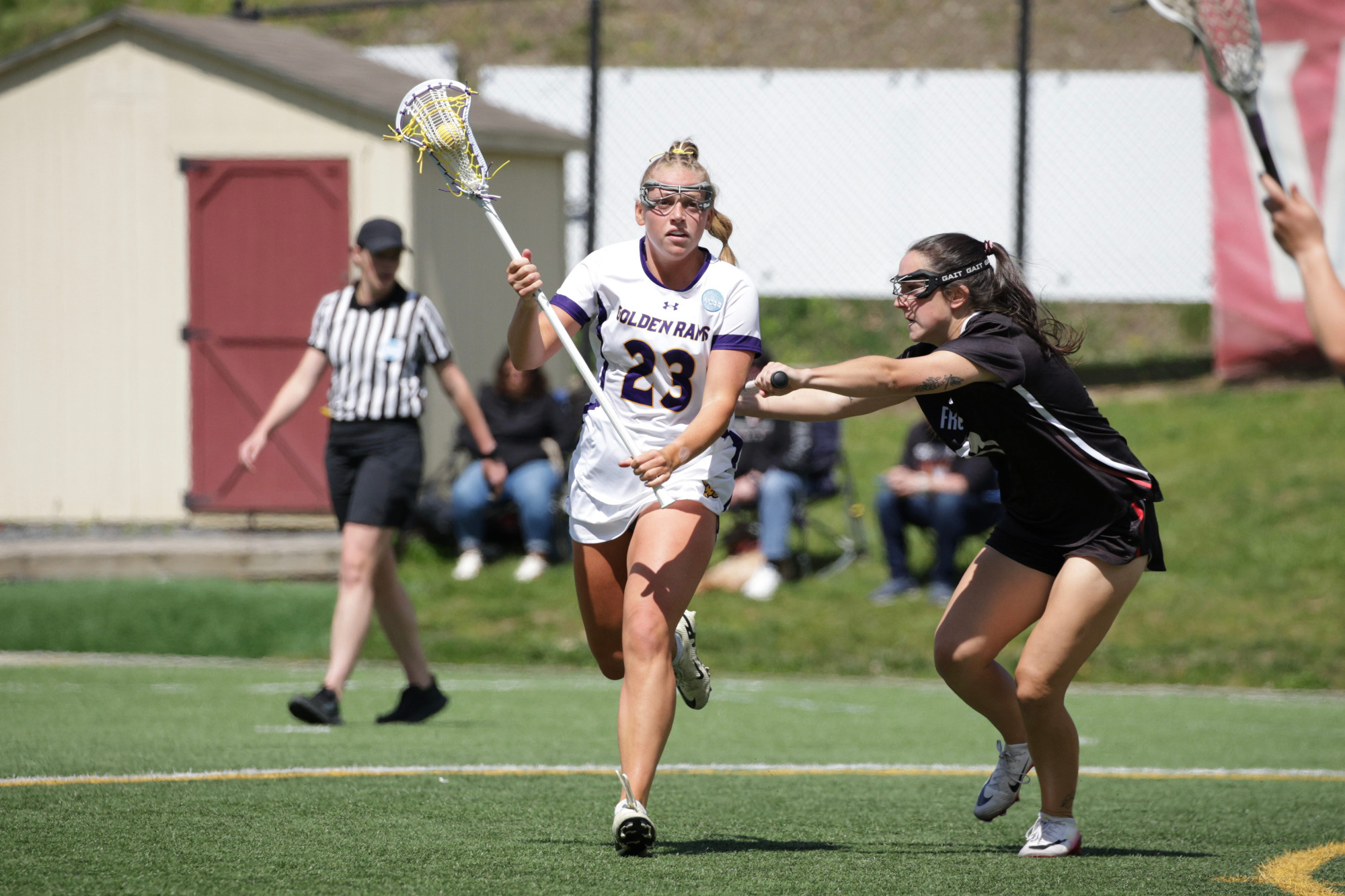 Lila McMahon-Skates works the ball around the top of the fan against a Frostburg State defender in the NCAA Tournament game at Whitenight Field in East Stroudsburg. Skates is wearing her home white jersey with purple numbers and purple trim while Frostburg State is wearing its road black jerseys with white numbers and orange trim.