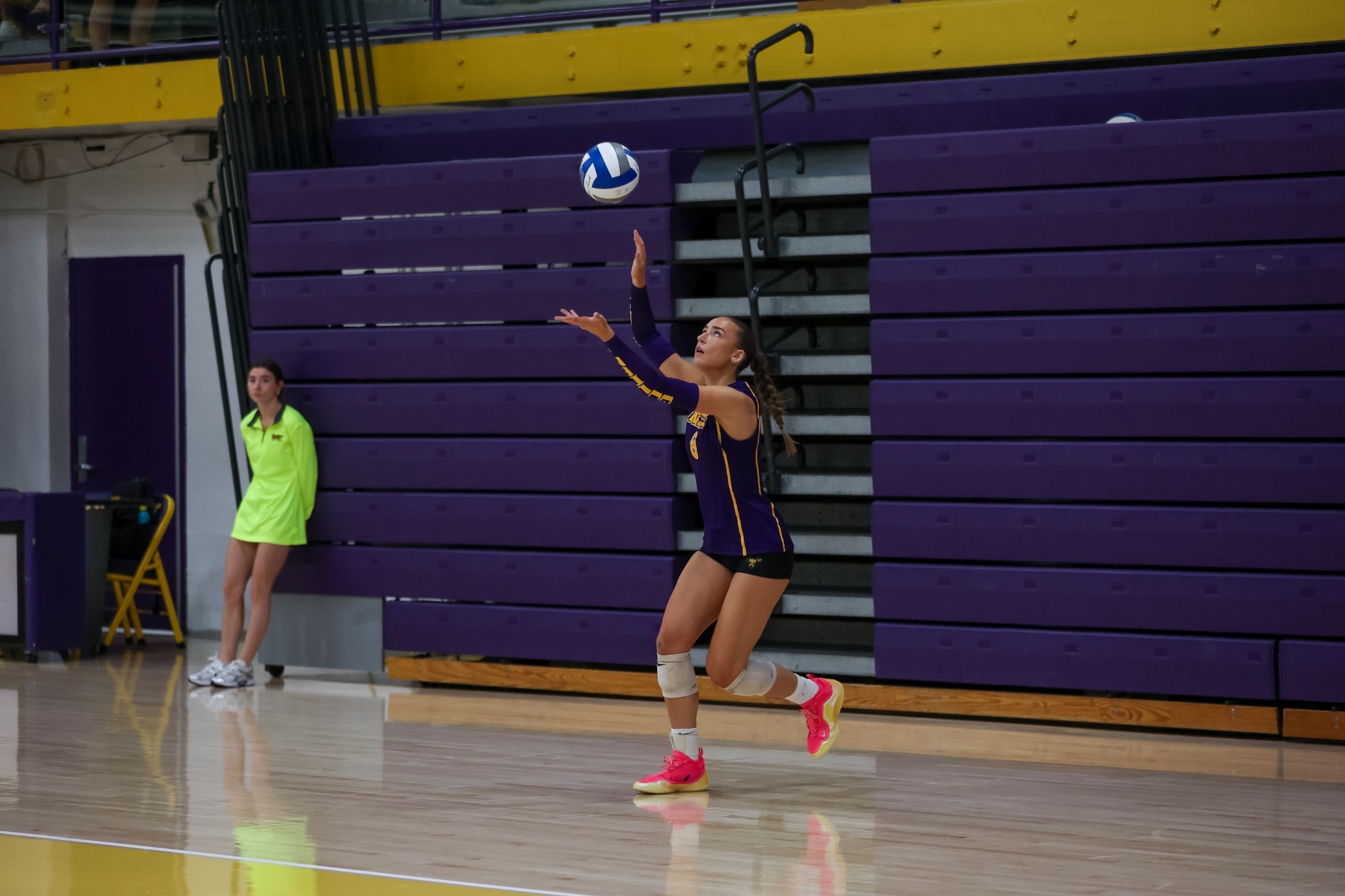 Brooke Forster throws the ball in the air to begin her serve vs Felician in the first set on Saturday afternoon in Hollinger. Forester wearing sleeveless purple and gold top with purple sleeves on forearms that say Golden Rams