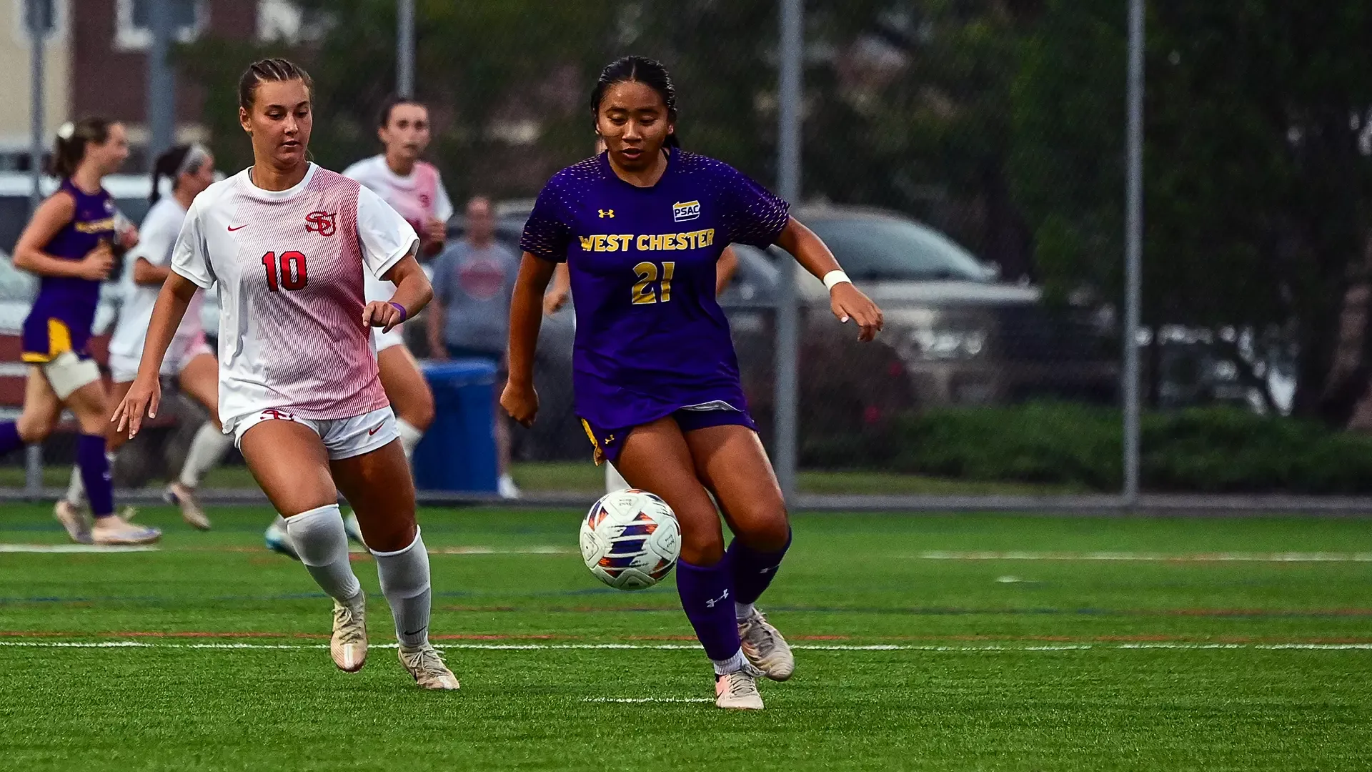 Jordan Nguyen wearing purple and gold road kits with her back to a Shippensburg player and the ball at her feet during Saturday night's match in Shippensburg
