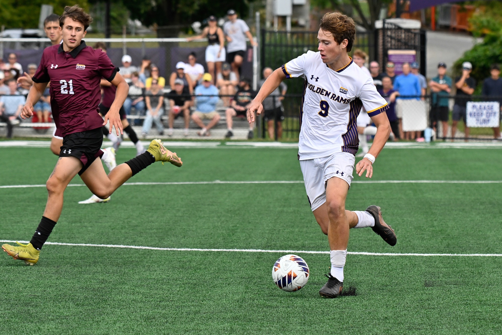 Keagan Penny wearing home white kit with number nine in purple on chest dribbles past a Bloomsburg defender wearing maroon top with black collar on Saturday afternoon