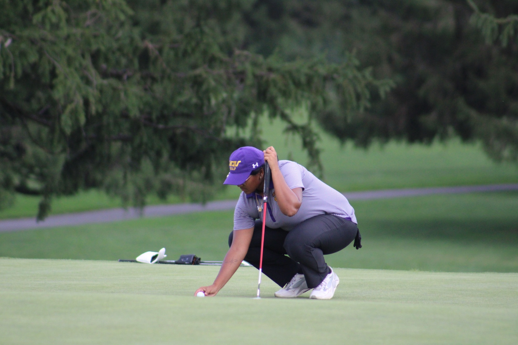 Hannah Alan wearing purple striped polo and black pants lines up a putt on the green with the golf ball in her right hand and putter in her left