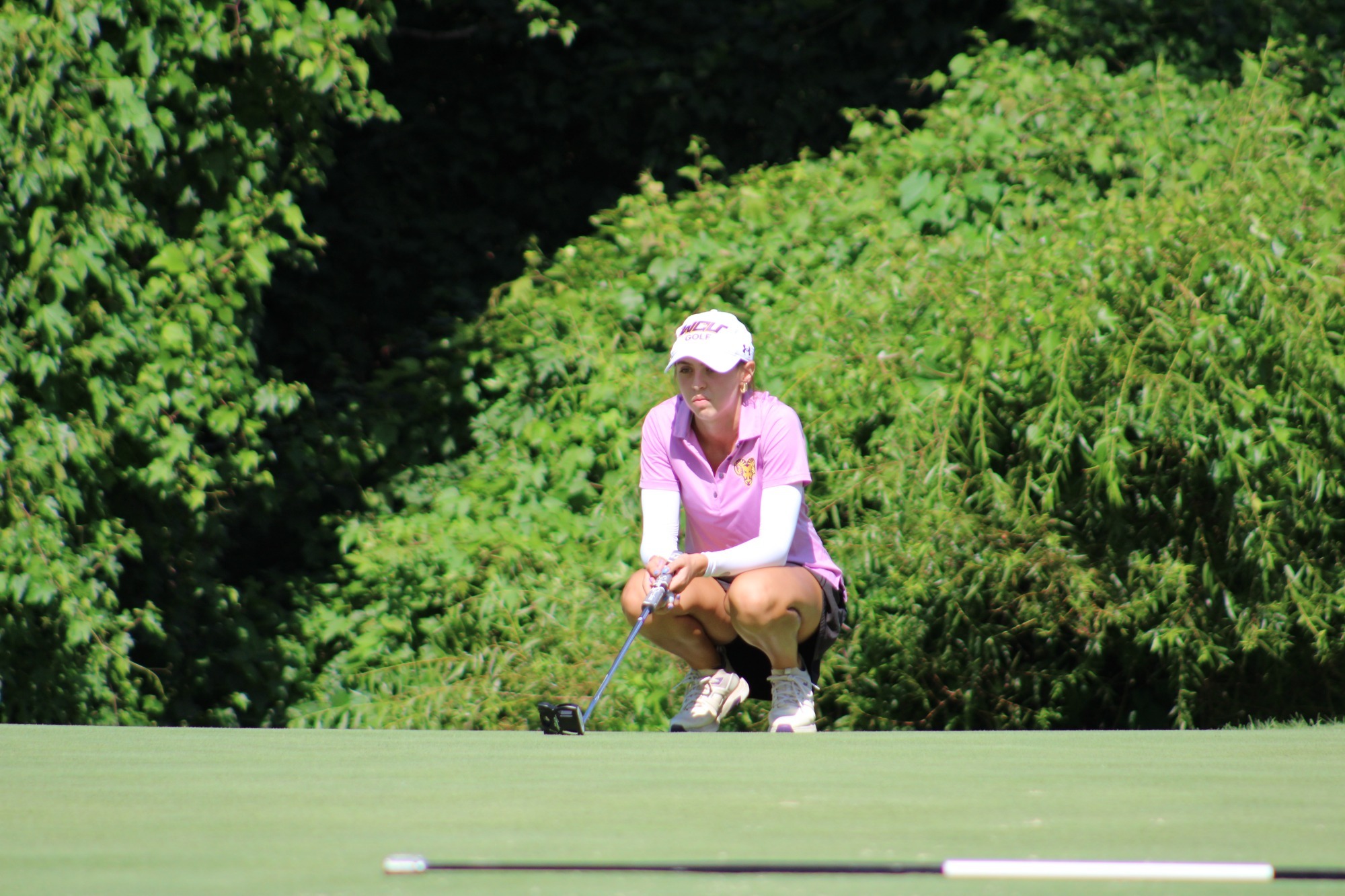 Emerson Glending wearing pink polo and white WCU golf hat crouches behind a putt to line it up
