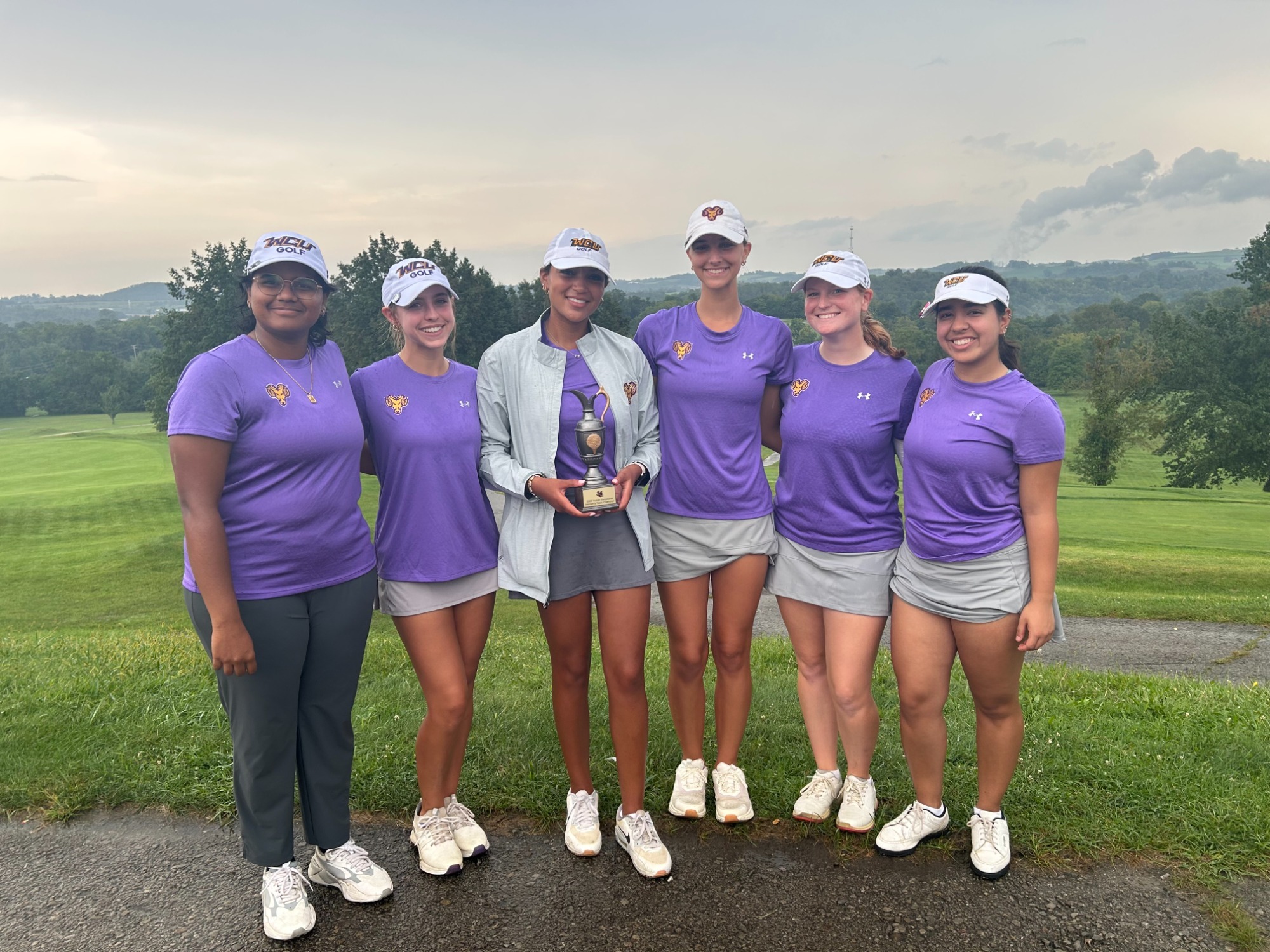 WOMEN'S GOLF TEAM POSING WITH VULCAN INVITATIONAL TEAM CHAMPIONSHIP TROPHY WEARING PURPLE TOPS AND GRAY BOTTOMS ON HILL OVERLOOKING GOLF COURSE