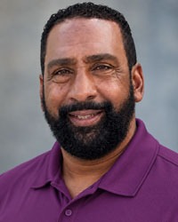 HEADSHOT TENNIS ASSISTANT COACH KEITH THOMPSON IN DARK PURPLE POLO INFRONT OF GRAY BACKGROUND