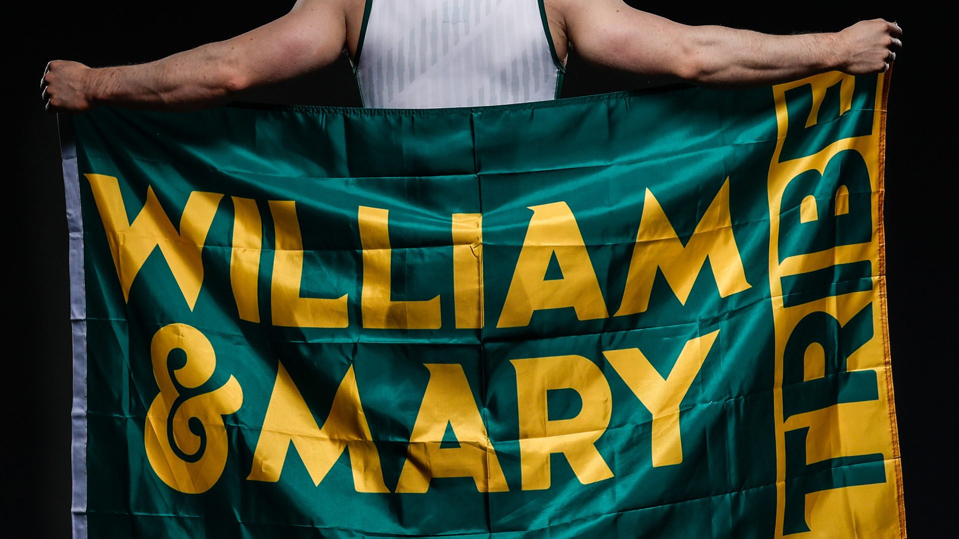 W&M men's gymnast holding a William & Mary Tribe flag with his back toward the camera